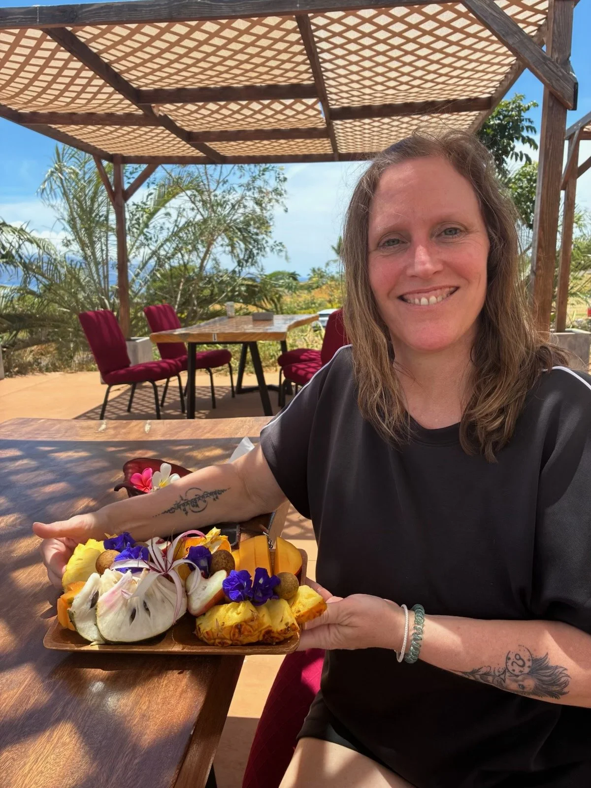 A woman is sitting at an outdoor wooden table under a thatched roof, smiling and holding a wooden platter of assorted tropical fruits decorated with edible flowers.