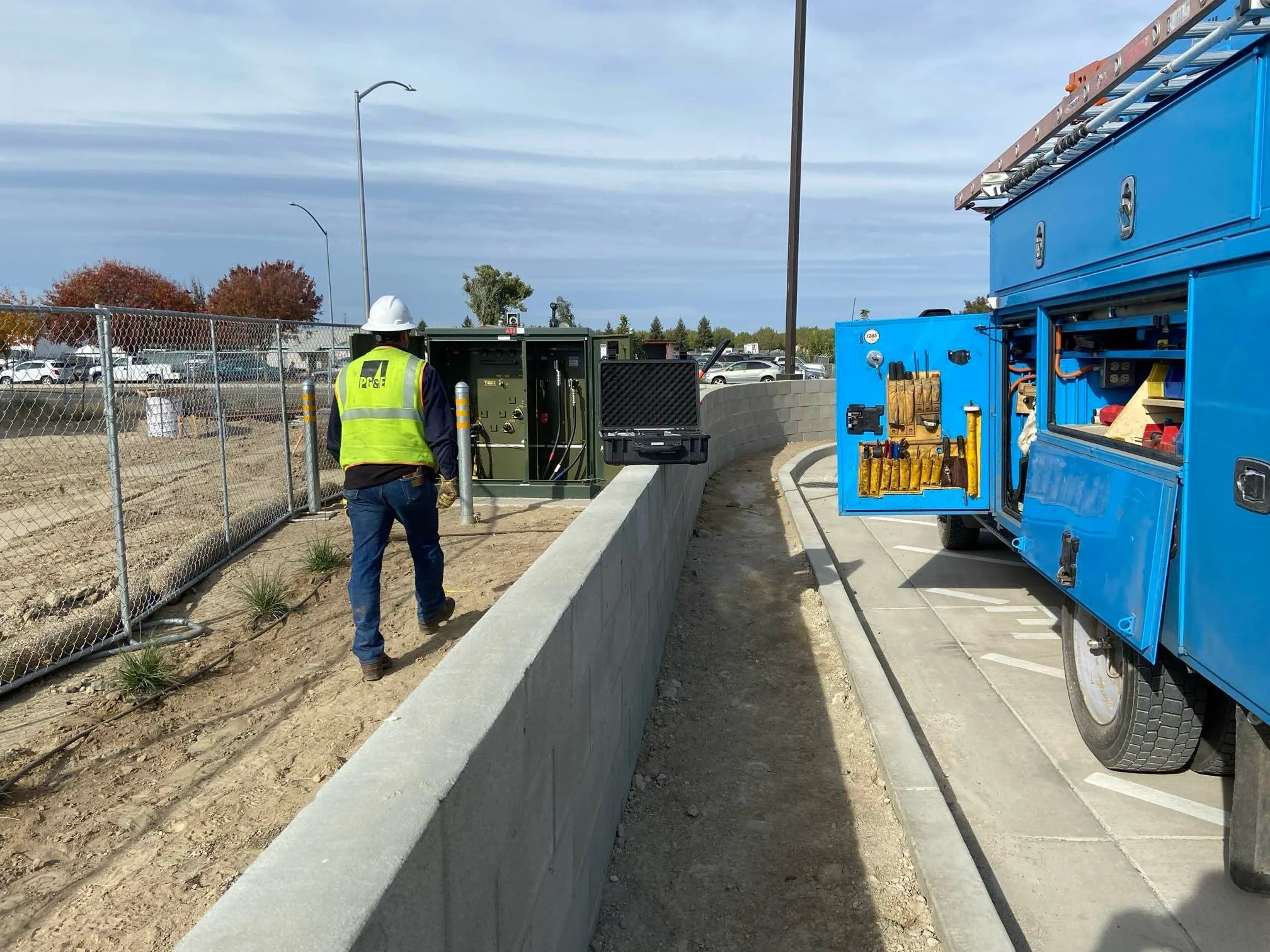 A construction worker wearing a safety vest and hard hat stands near electrical equipment installed on a low concrete wall, with a blue utility truck and construction tools nearby. There is a chain-link fence and parked cars in the background on a clear day.