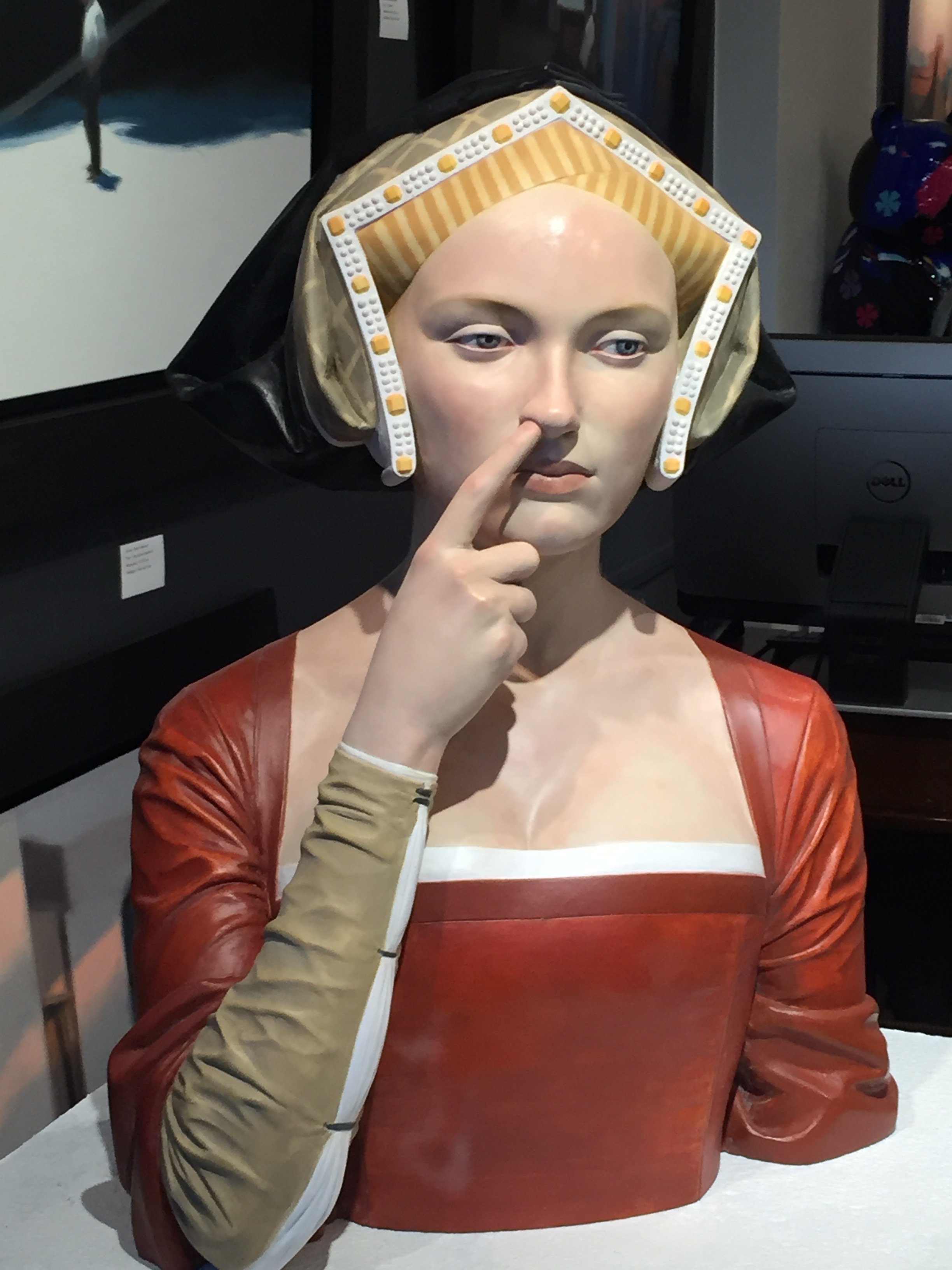A life-sized sculpture of a woman with a thoughtful expression, wearing a red dress with an off-shoulder design, and a beige arm sleeve, is displayed indoors.