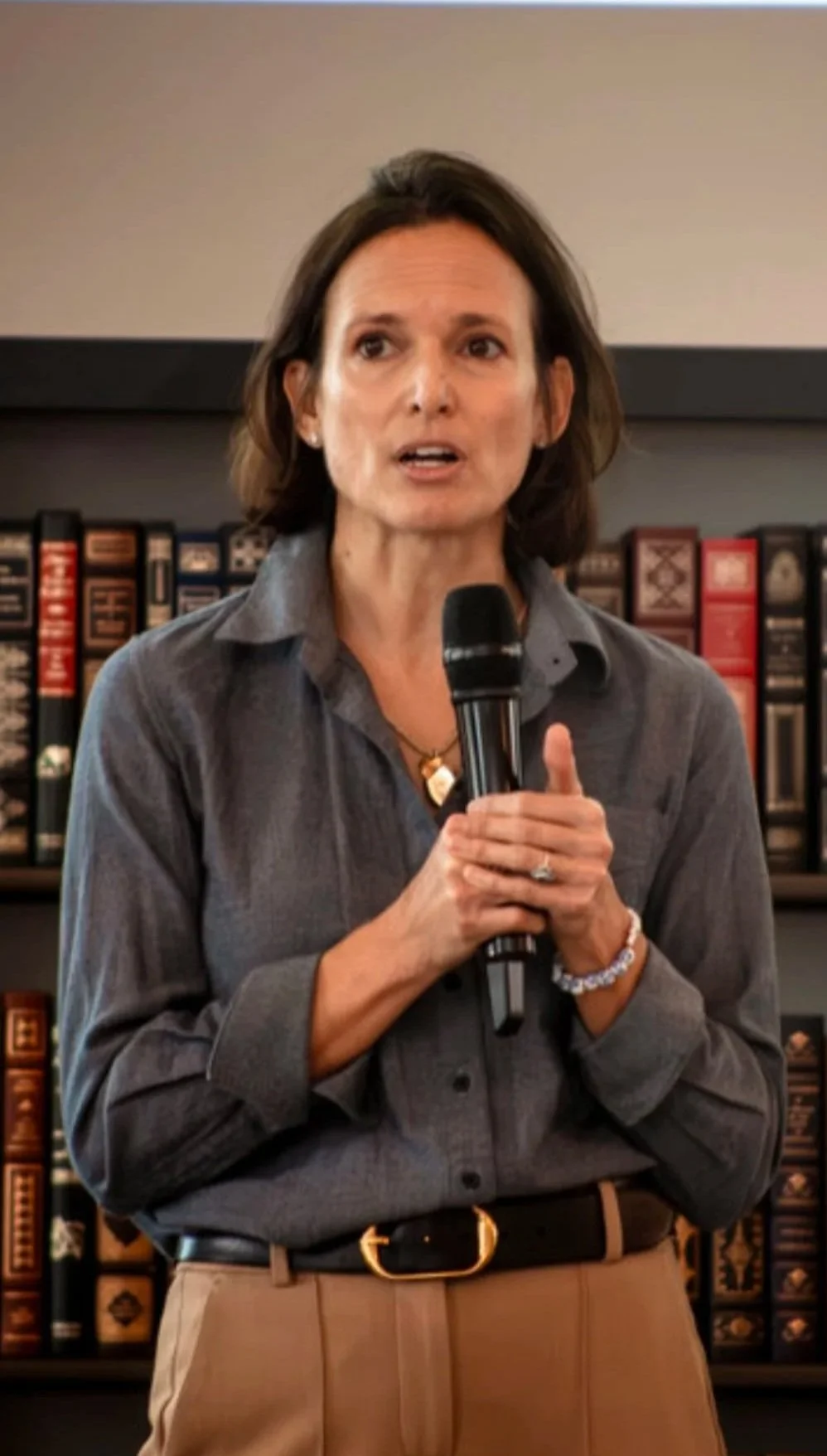 A woman holding a microphone and speaking, standing in front of a bookshelf filled with books.