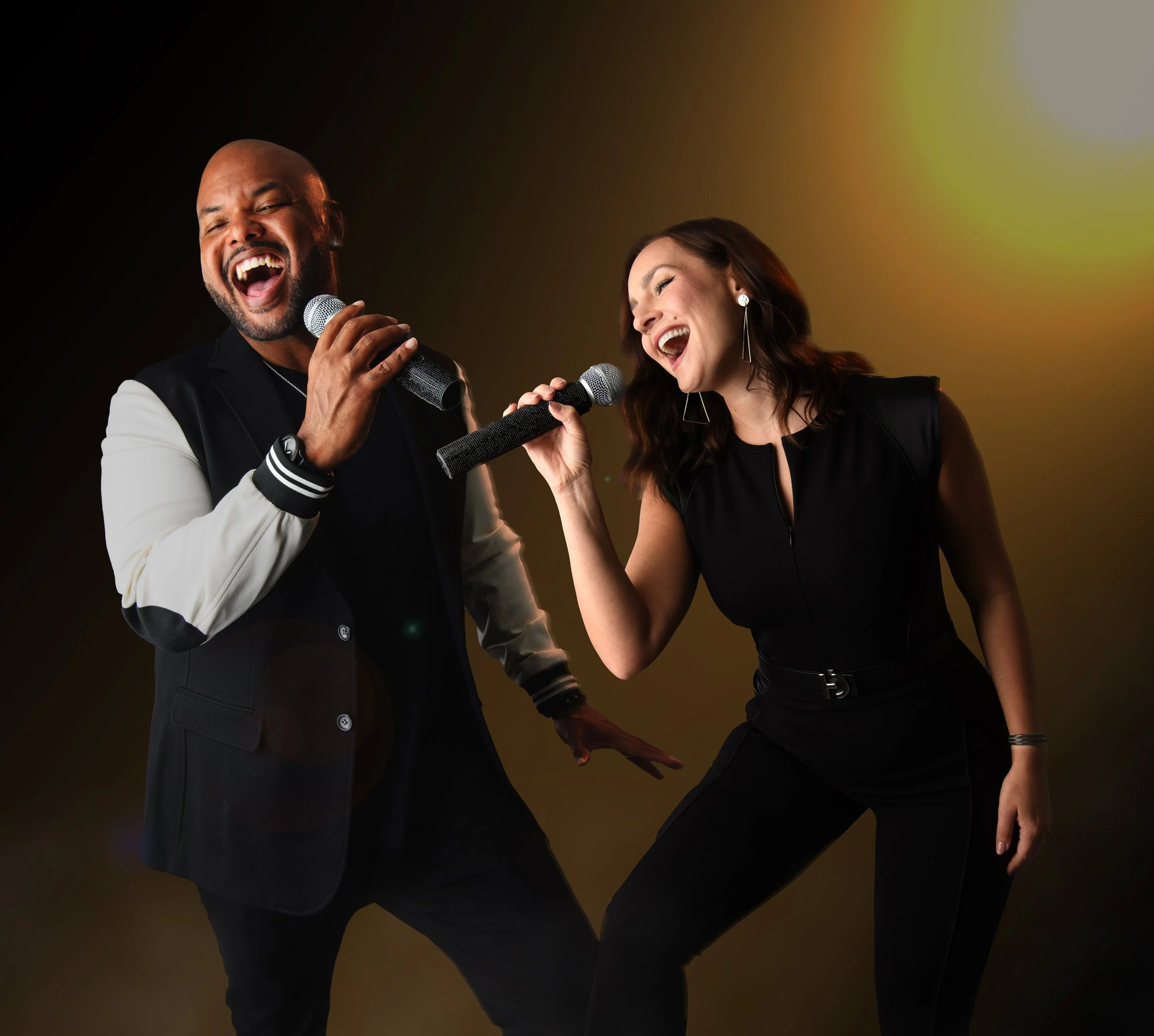 A man and a woman are singing into microphones, smiling and enjoying themselves. The man is bald with a beard and is wearing a varsity jacket, while the woman has long brown hair and is wearing earrings and a black top. The background is a gradient of dark brown, yellow, and white.