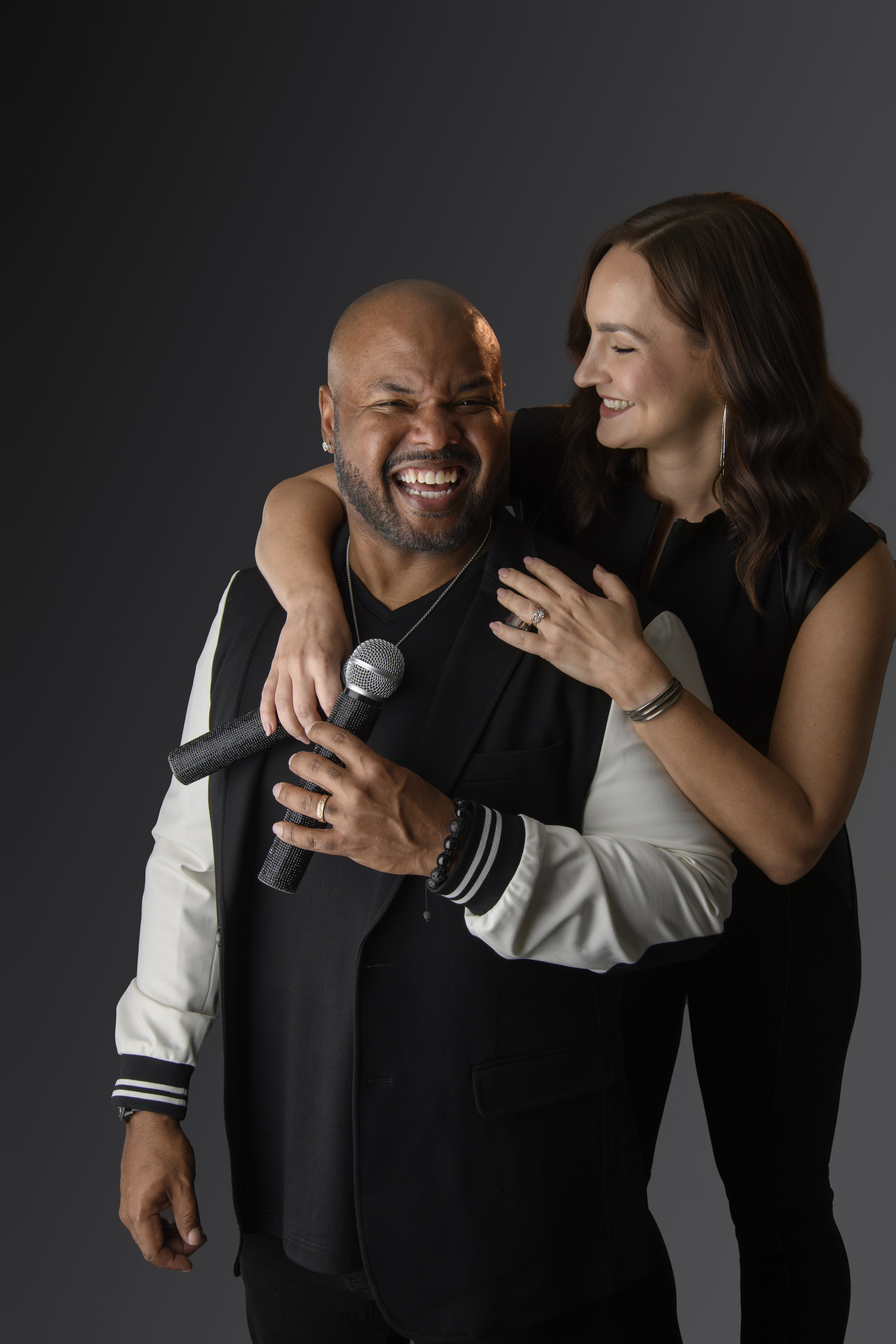 A man and woman sharing a joyful moment, with the woman leaning on the man's shoulder. The man is holding a microphone and wearing a black and white jacket. The woman has dark wavy hair, smiling, with her arm around the man's shoulders and her hand on his chest. The background is a plain dark gray.