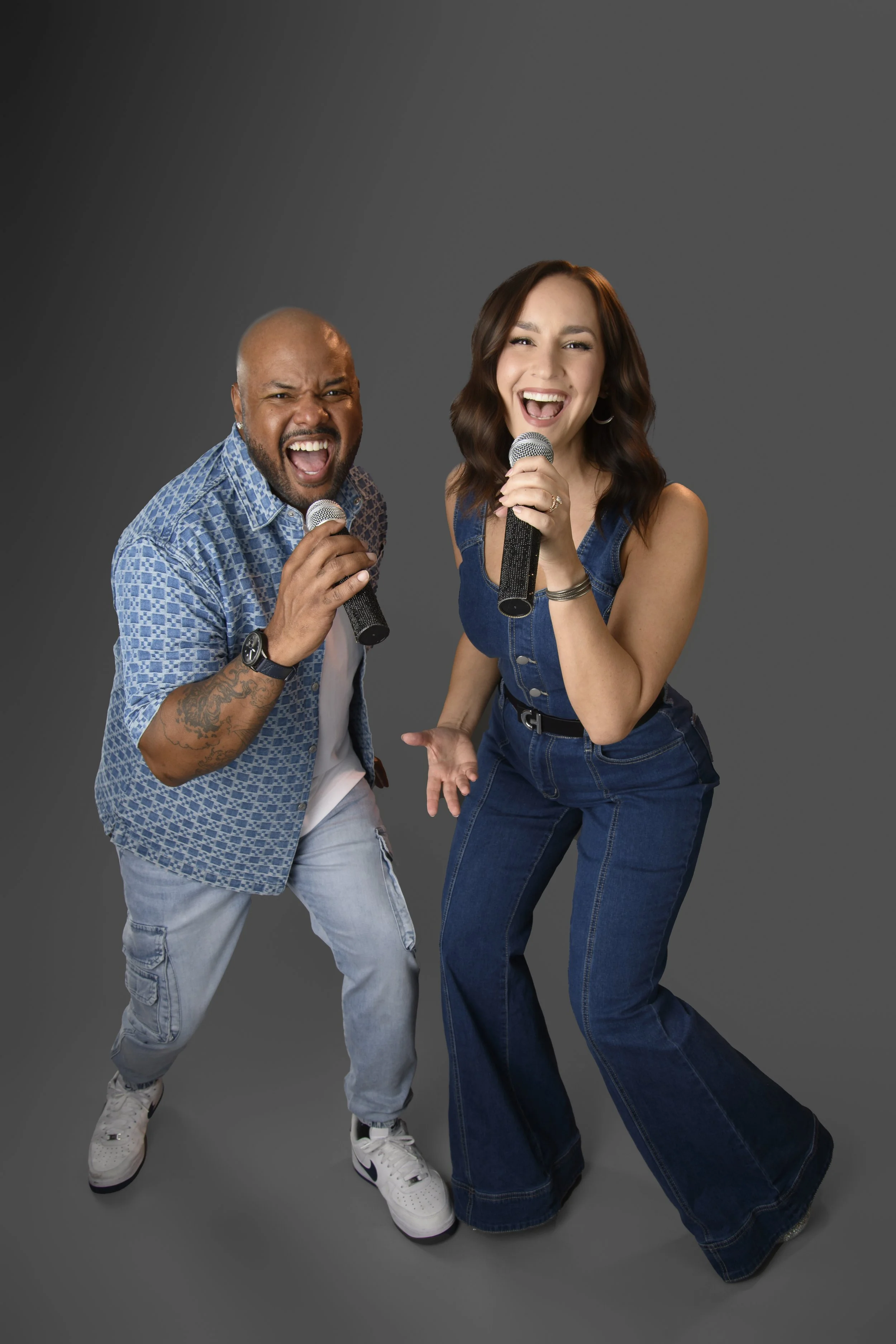 Two people, a man and a woman, singing into microphones and looking joyful. The man is bald, with a beard, wearing a blue patterned short-sleeve shirt and light blue jeans, while the woman has long wavy brown hair, wearing a sleeveless denim top and matching pants.