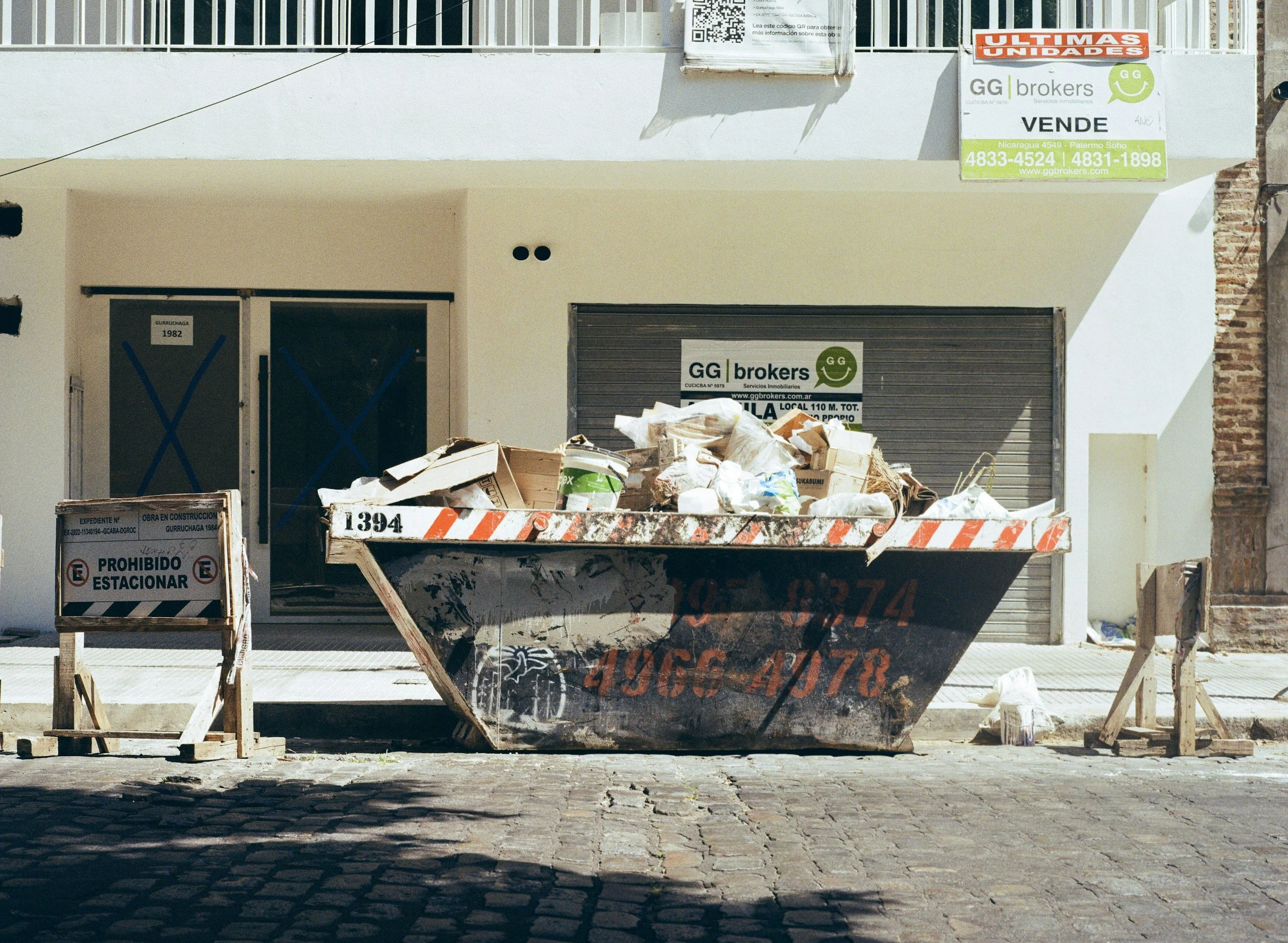 Construction site featuring a rusty container truck filled with construction debris and a 'no parking' sign in Spanish.