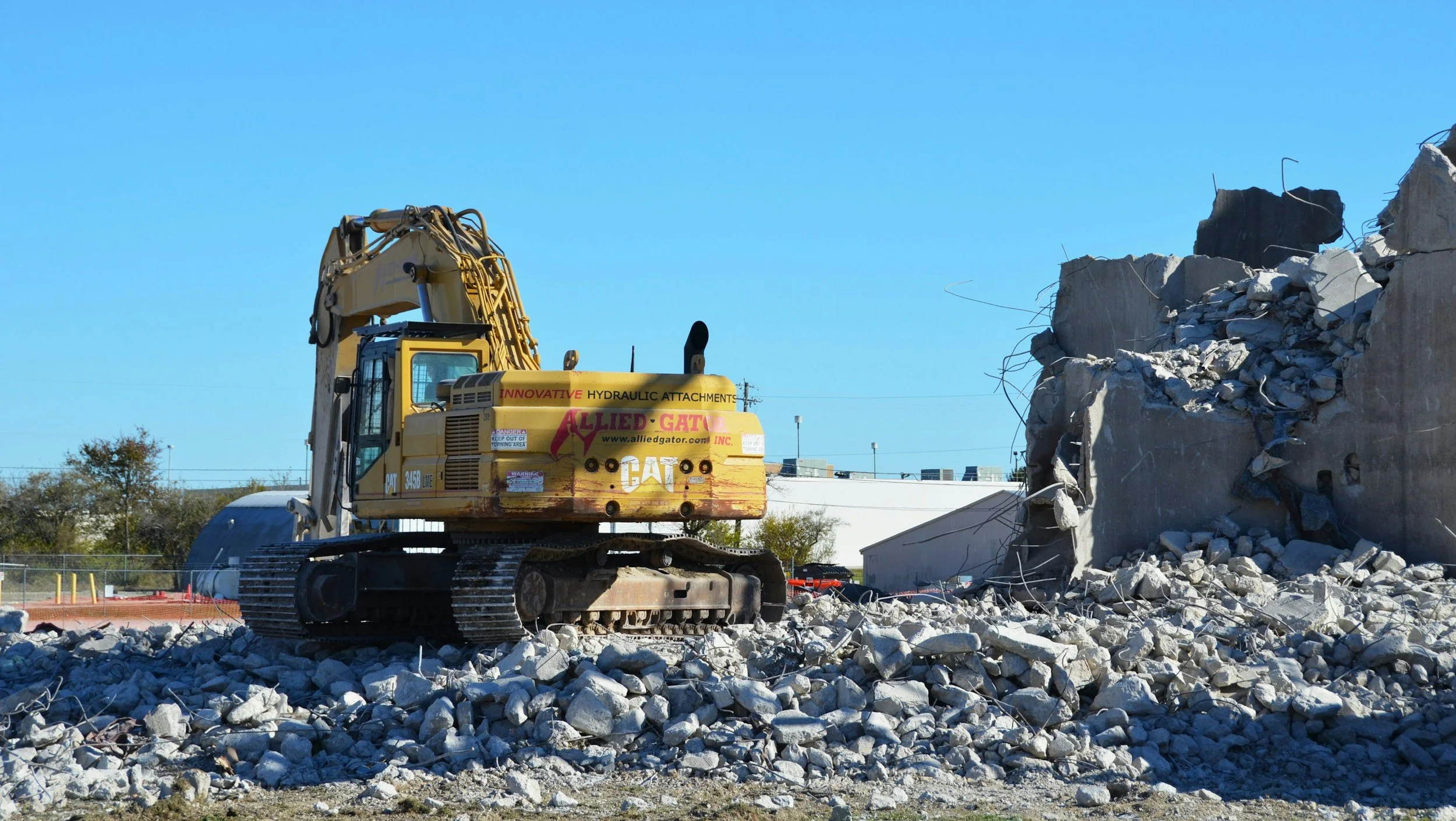 A yellow excavator working on demolishing a concrete building, with debris and rubble around.