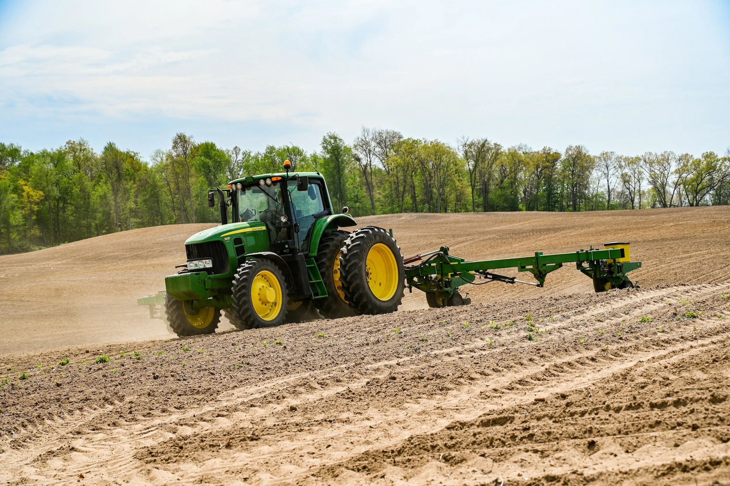 A green tractor working in a field with tilled soil, surrounded by trees and a cloudy sky.