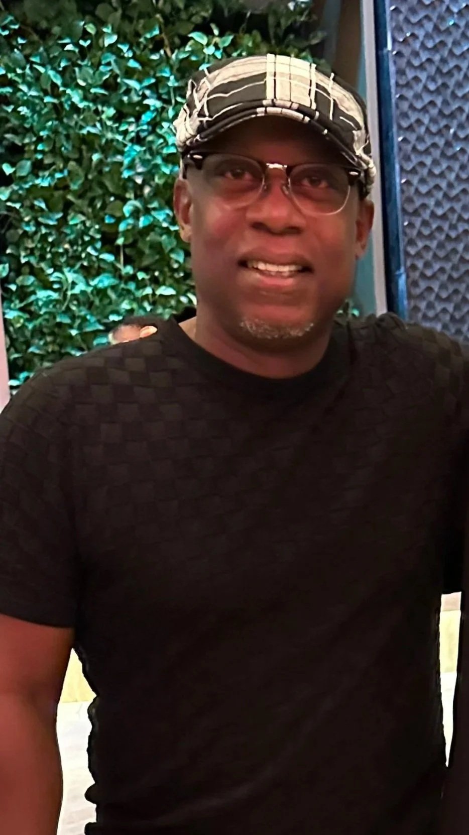 A man smiling, wearing glasses, a striped cap, and a dark patterned shirt. Behind him is a green leafy background and a textured blue wall.