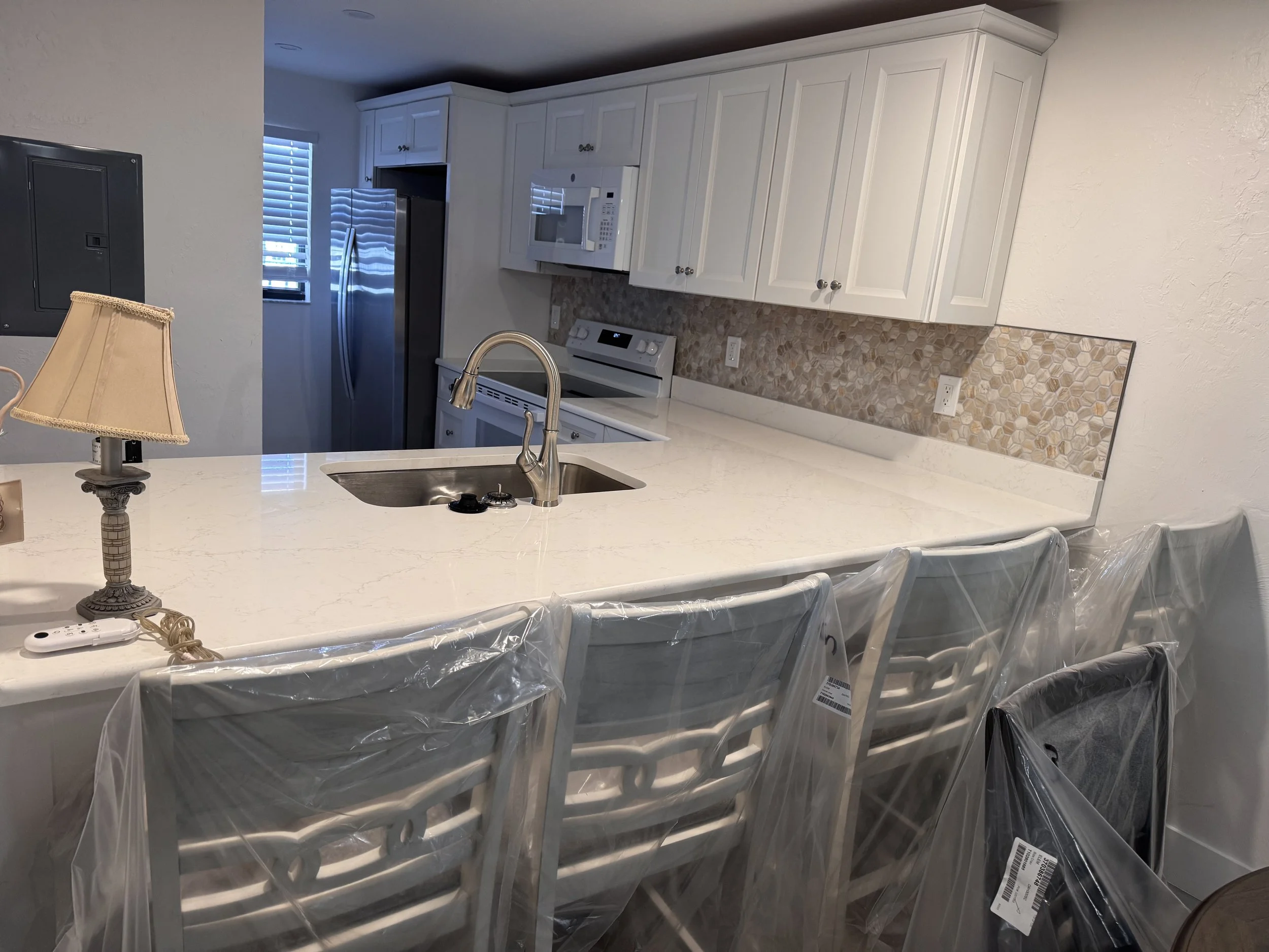 A kitchen with white cabinets, a stainless steel refrigerator, a microwave oven, a stove, a beige tile backsplash, and a white countertop. There is a small table lamp and chairs covered with plastic.