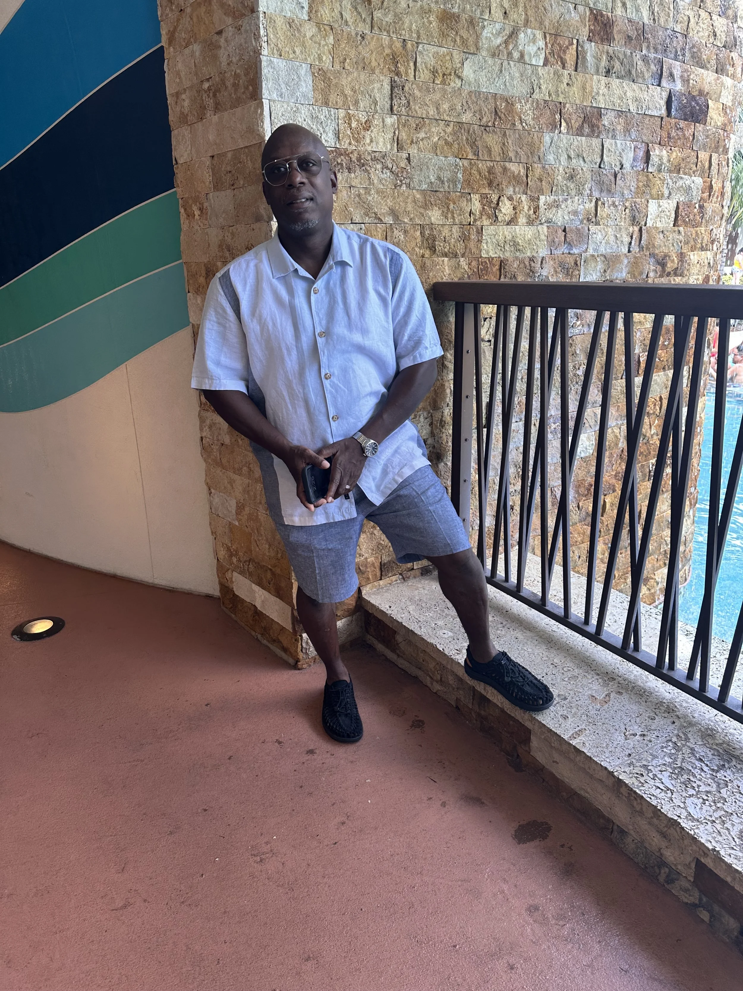 A man standing next to a stone wall, wearing a light blue shirt, shorts, glasses, and black sneakers, holding a phone in one hand, with a railing on his right and a pool visible outside.