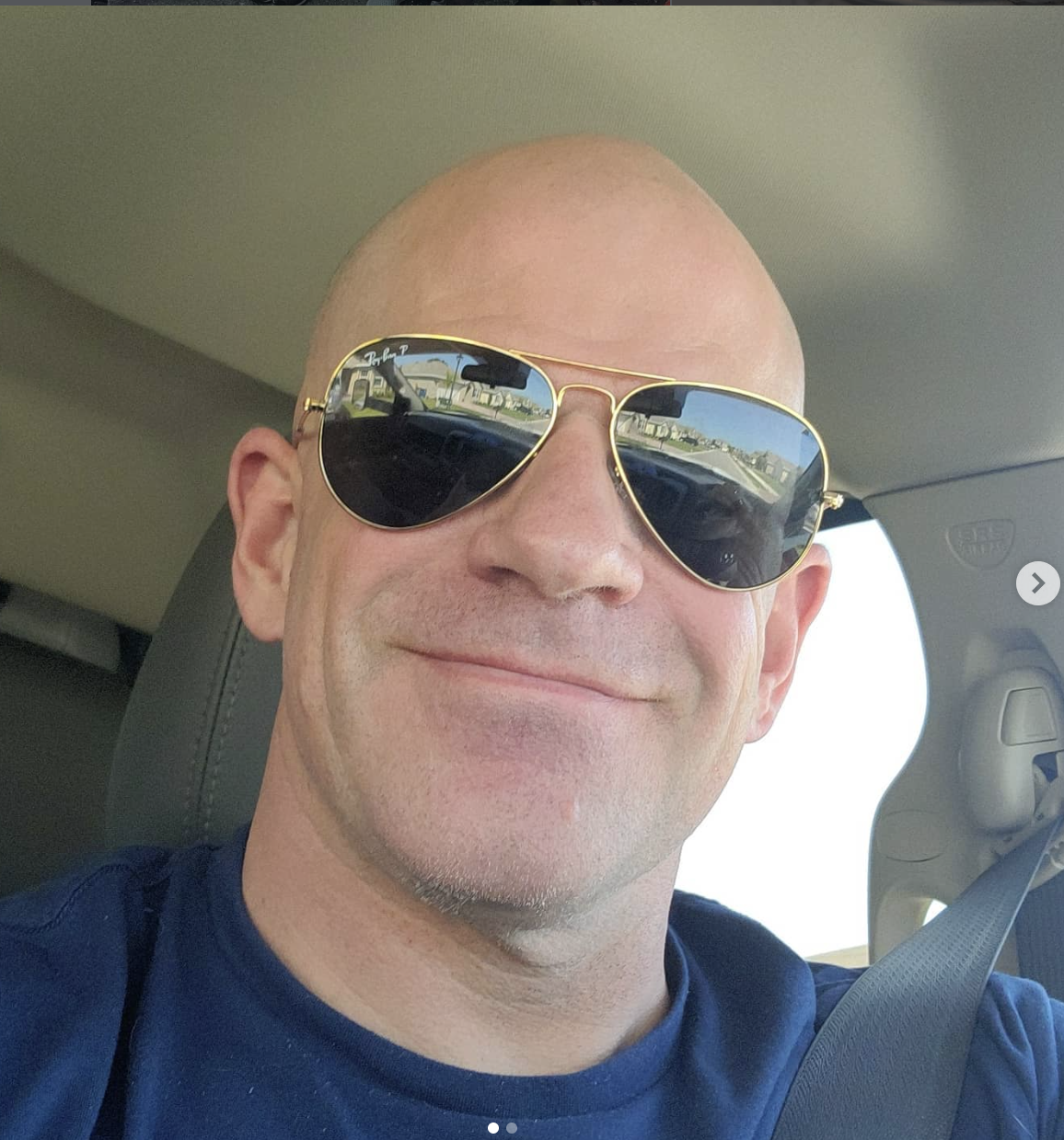 A man with a bald head, wearing sunglasses and a blue shirt, sitting in a car and smiling.
