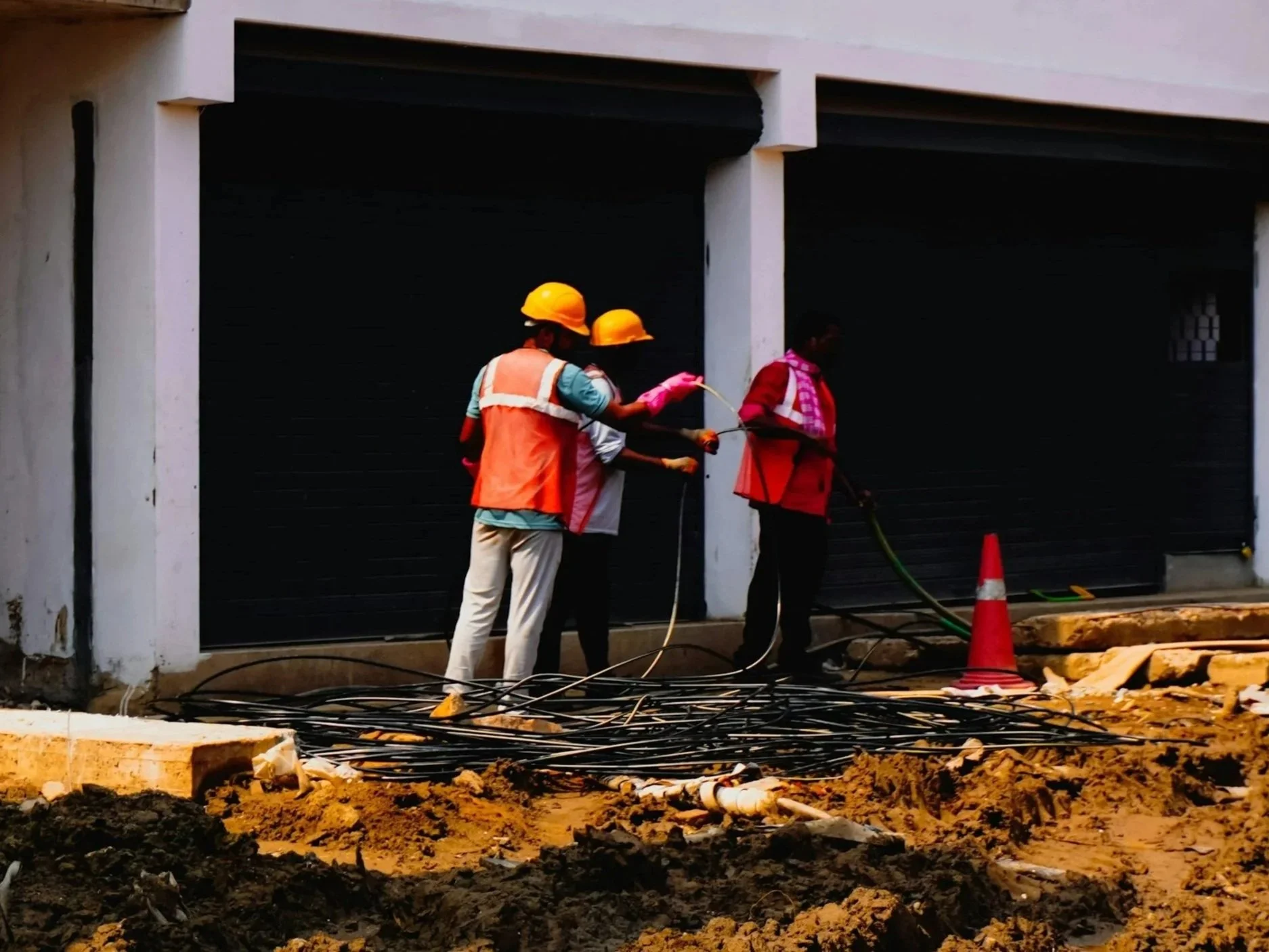 Construction workers wearing yellow helmets and vests working on wiring in front of a large black door, with construction cones and dirt ground in the foreground.