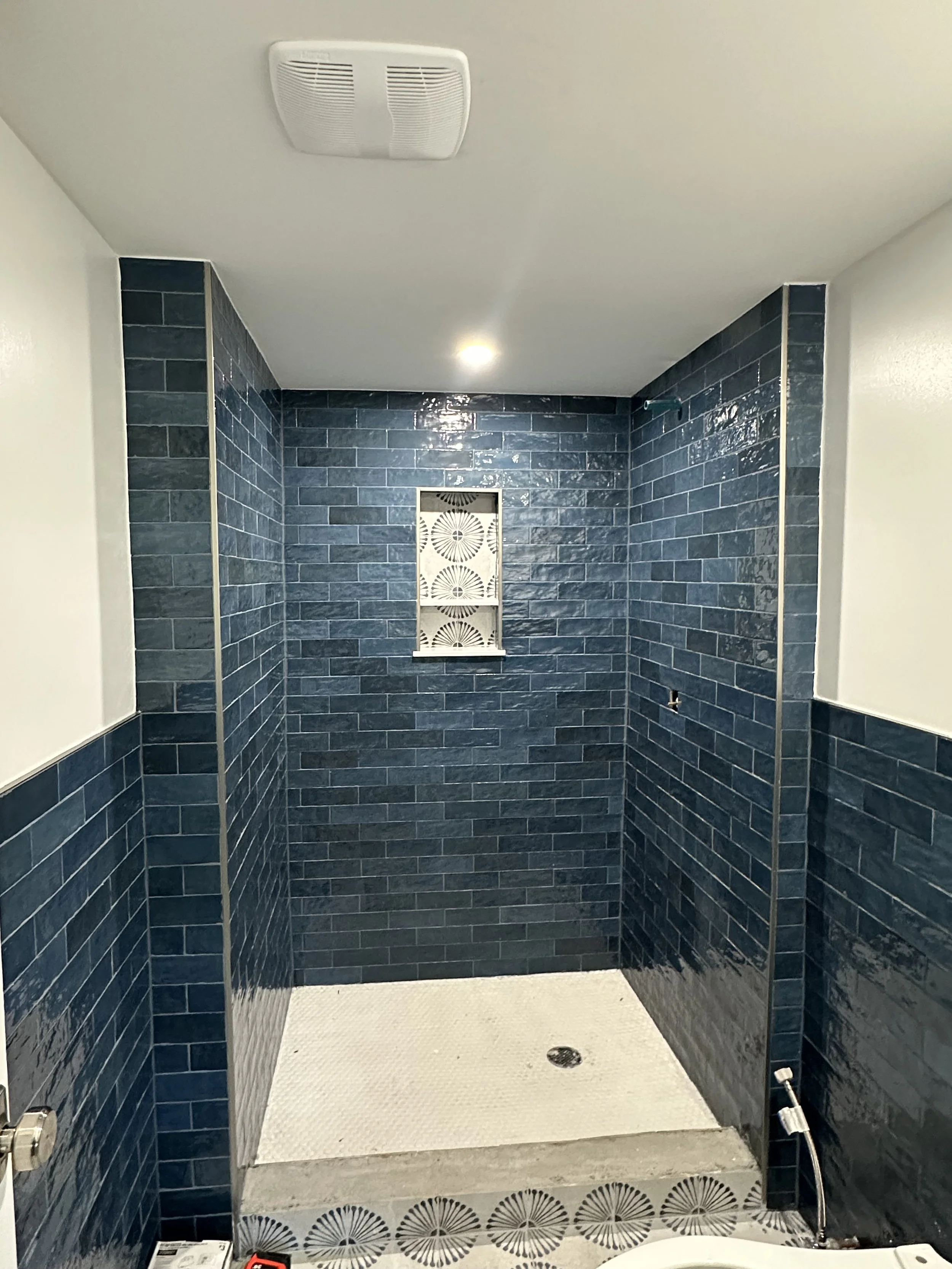 Shower stall with blue subway tile walls, white textured floor, a small window with decorative grille, and a ceiling vent.