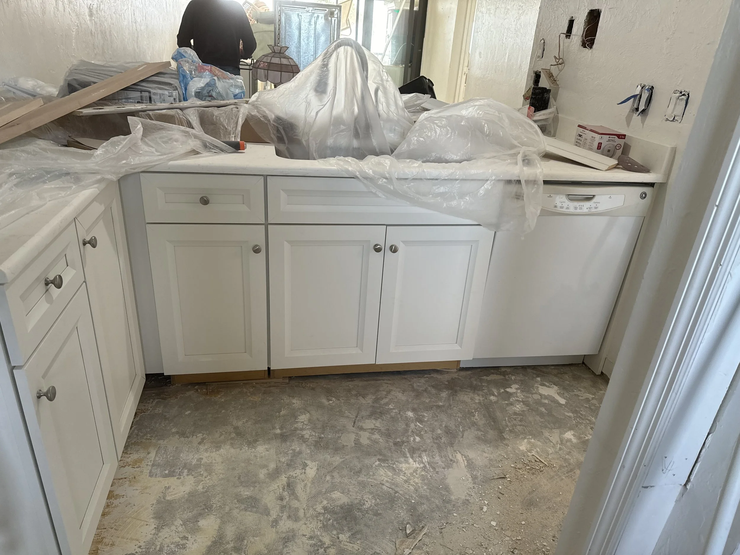 A kitchen under renovation with white cabinets, some plastic sheets, construction materials, and an unfinished floor.