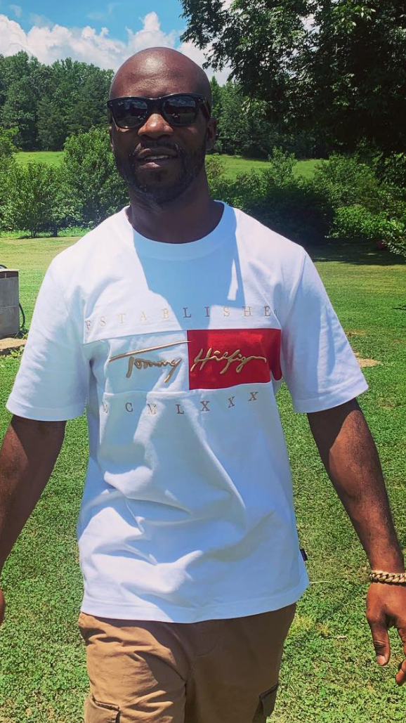 A man standing outdoors on a sunny day, wearing sunglasses, a white Tommy Hilfiger t-shirt, and tan pants. He has a dark complexion and a beard, with trees and a grassy area in the background.