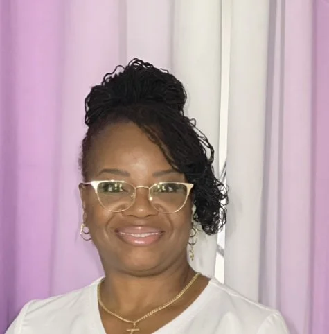 Portrait of a smiling woman with glasses, jewelry, and braided hair styled in an updo, standing in front of purple and white curtains.