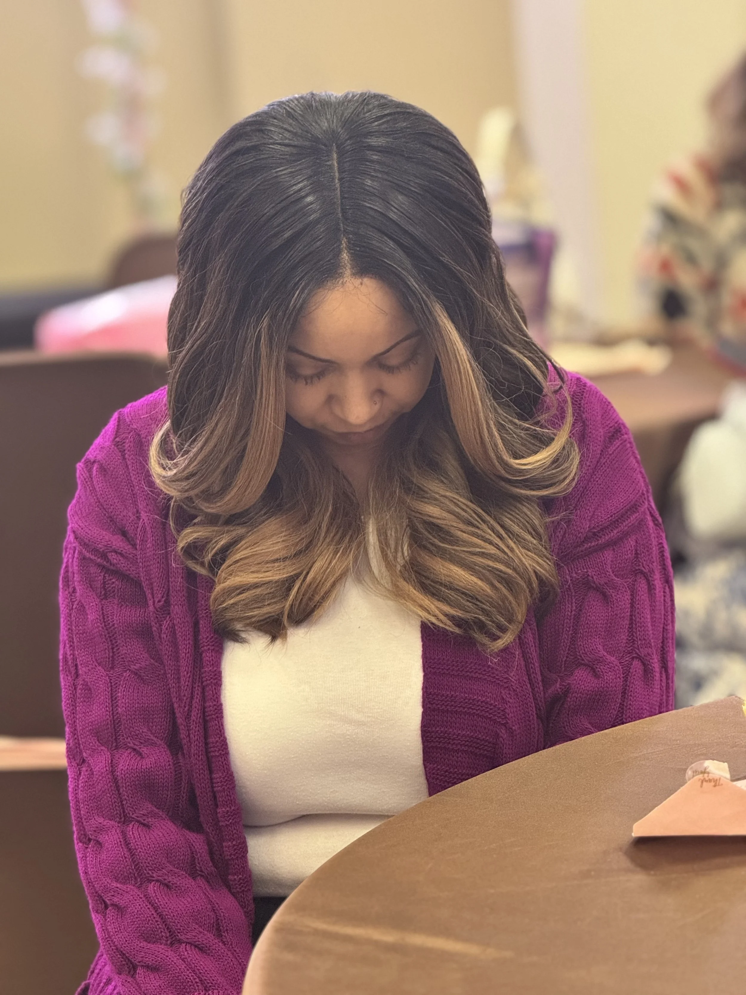 Woman with long, wavy hair wearing a purple sweater and white top, looking downward, sitting at a table in a room with a blurred background.