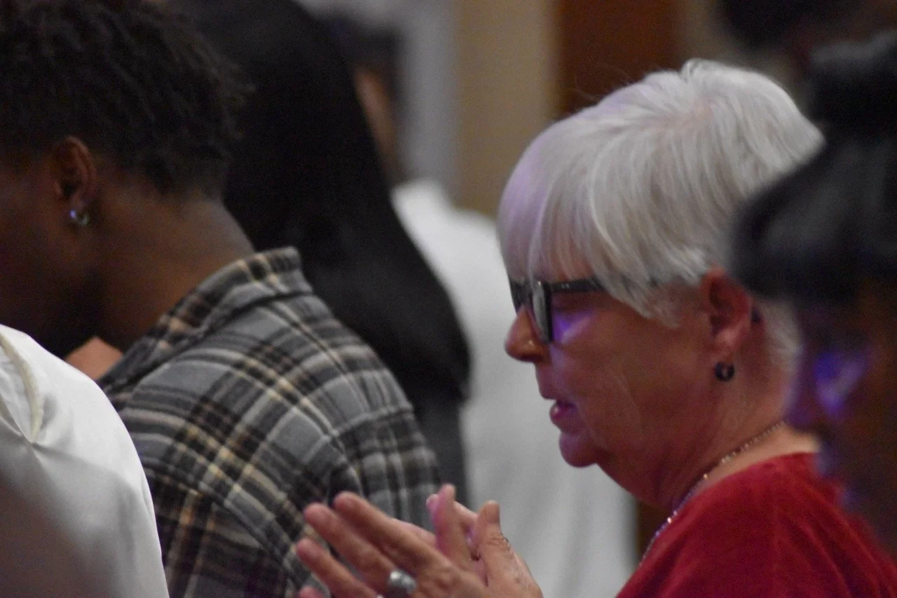 An elderly white woman with white hair, wearing glasses and a red top, is praying or engaging in a moment of reflection with her hands clasped.