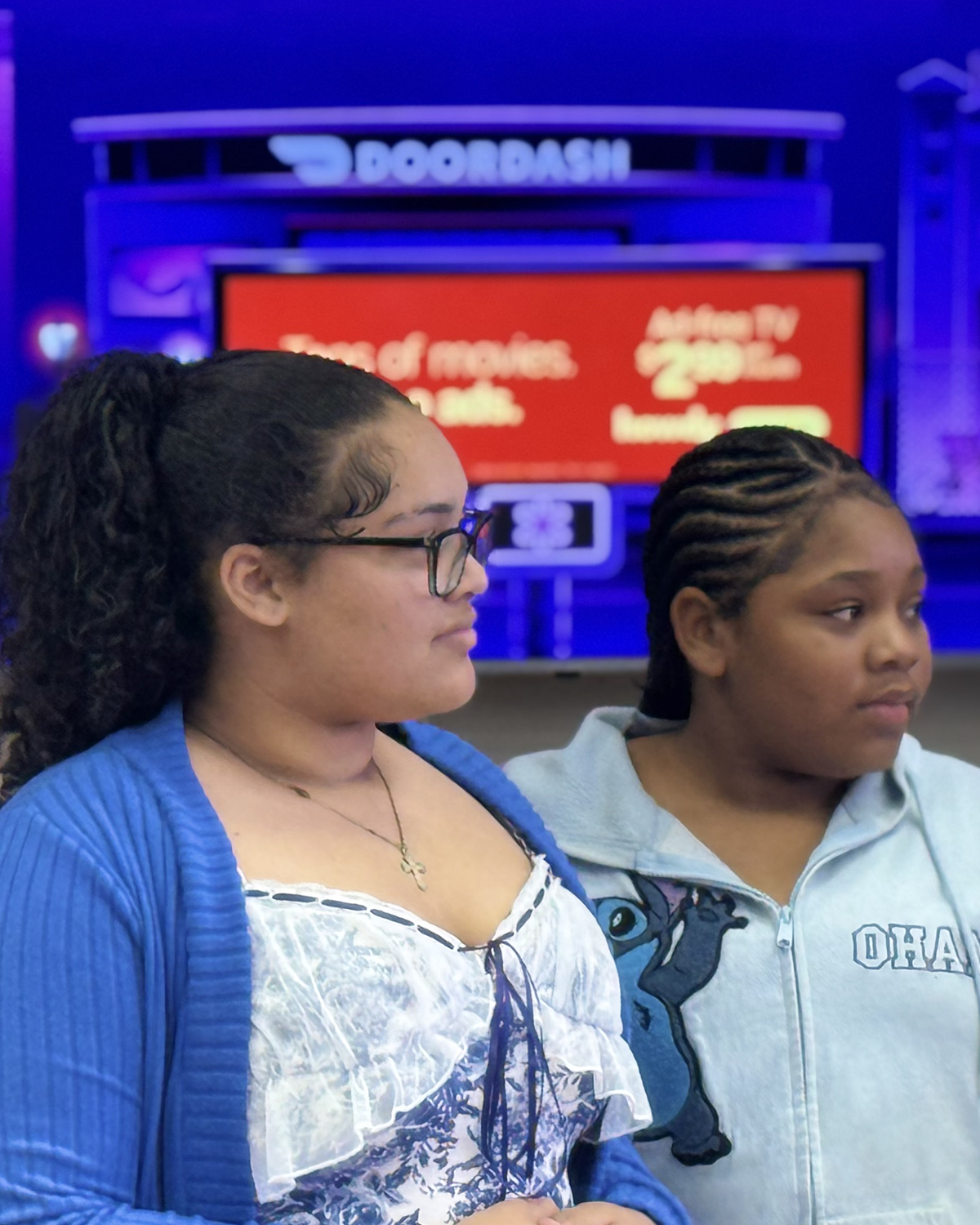 Two young women standing side by side indoors, one with glasses and long dark hair in a ponytail, wearing a blue cardigan over a white blouse with ruffles and a necklace. The other with braided hair, wearing a light gray hoodie with a horse graphic and the word "OHA". In the background, a red digital screen and a blue neon sign reading "D oorDash" are visible.