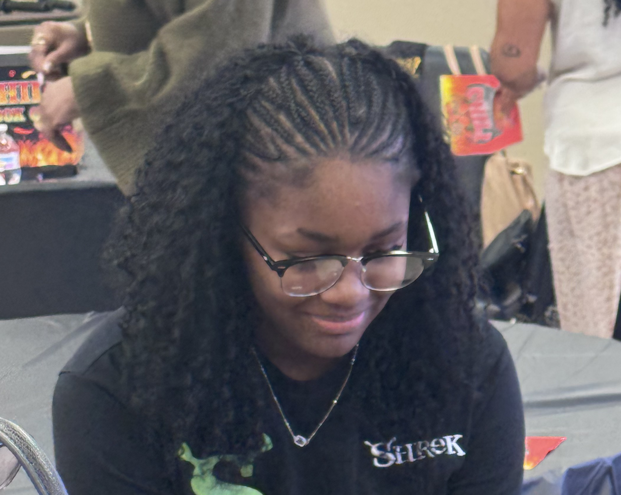 A young woman with curly hair and glasses sitting at a table, looking down and smiling.