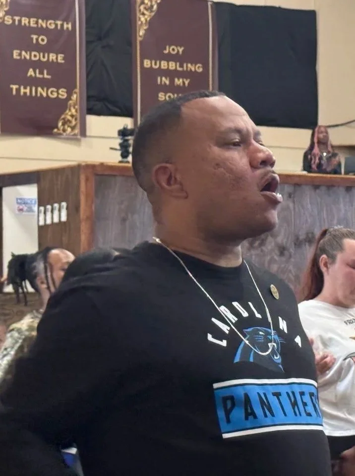 Man singing or speaking passionately during a gathering or event, wearing a Carolina Panthers T-shirt, with people in the background and motivational banners on the wall.