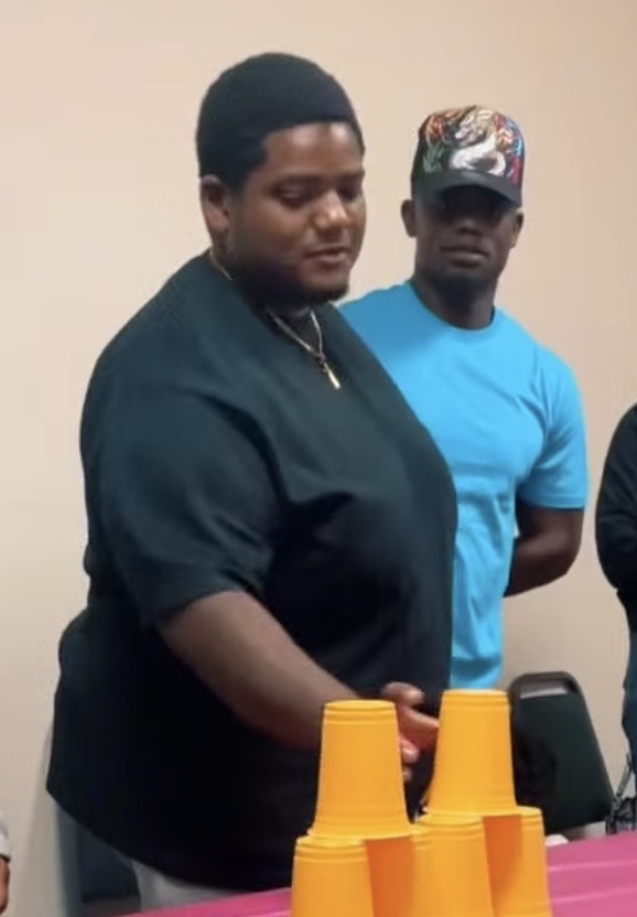 Two men standing behind table with stacked yellow cups, in a room with plain beige walls.