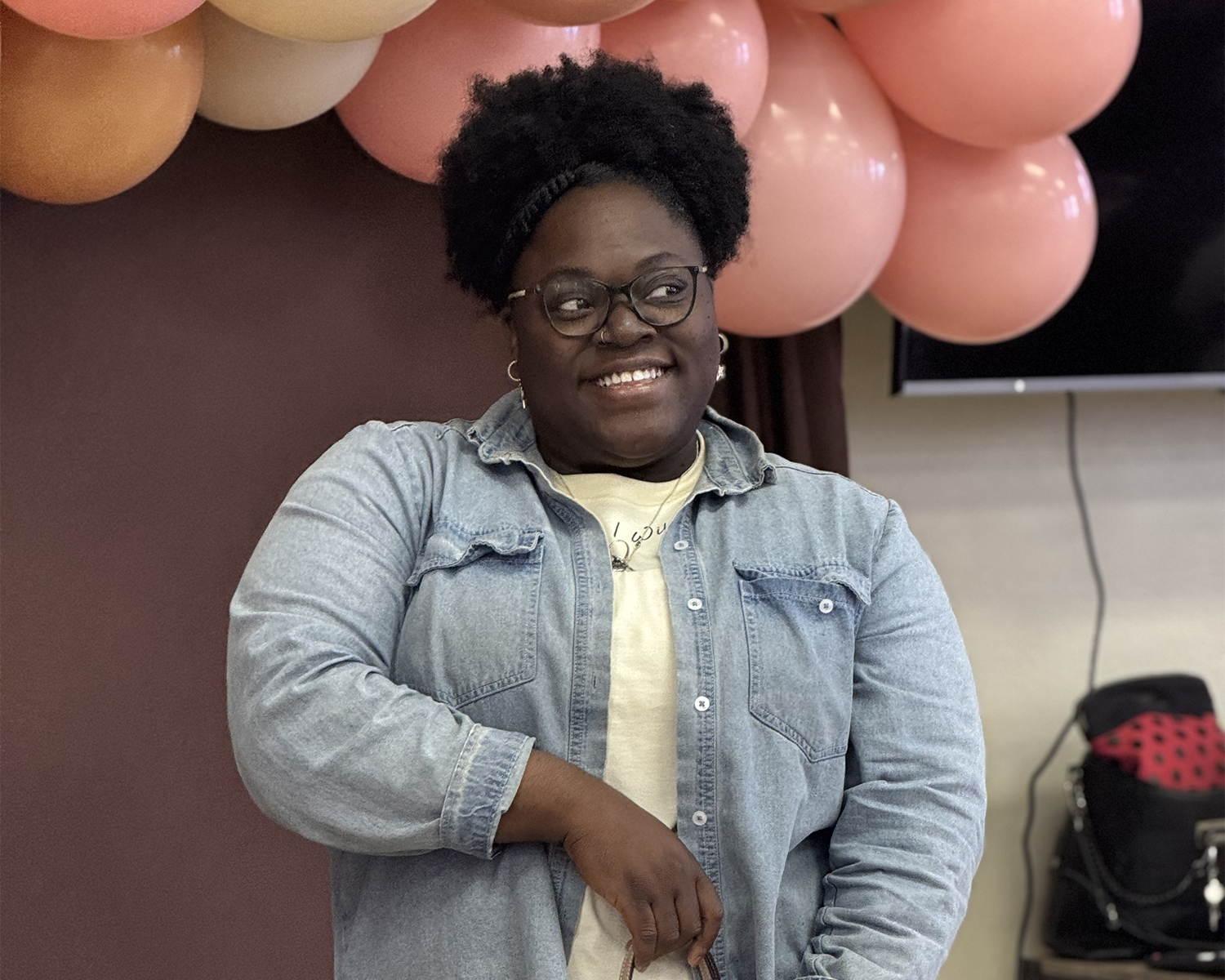 A woman with dark skin and glasses, smiling, standing in front of pink balloons. She is wearing a denim jacket over a light-colored shirt, with her hand resting on her arm. A bag is visible in the background.