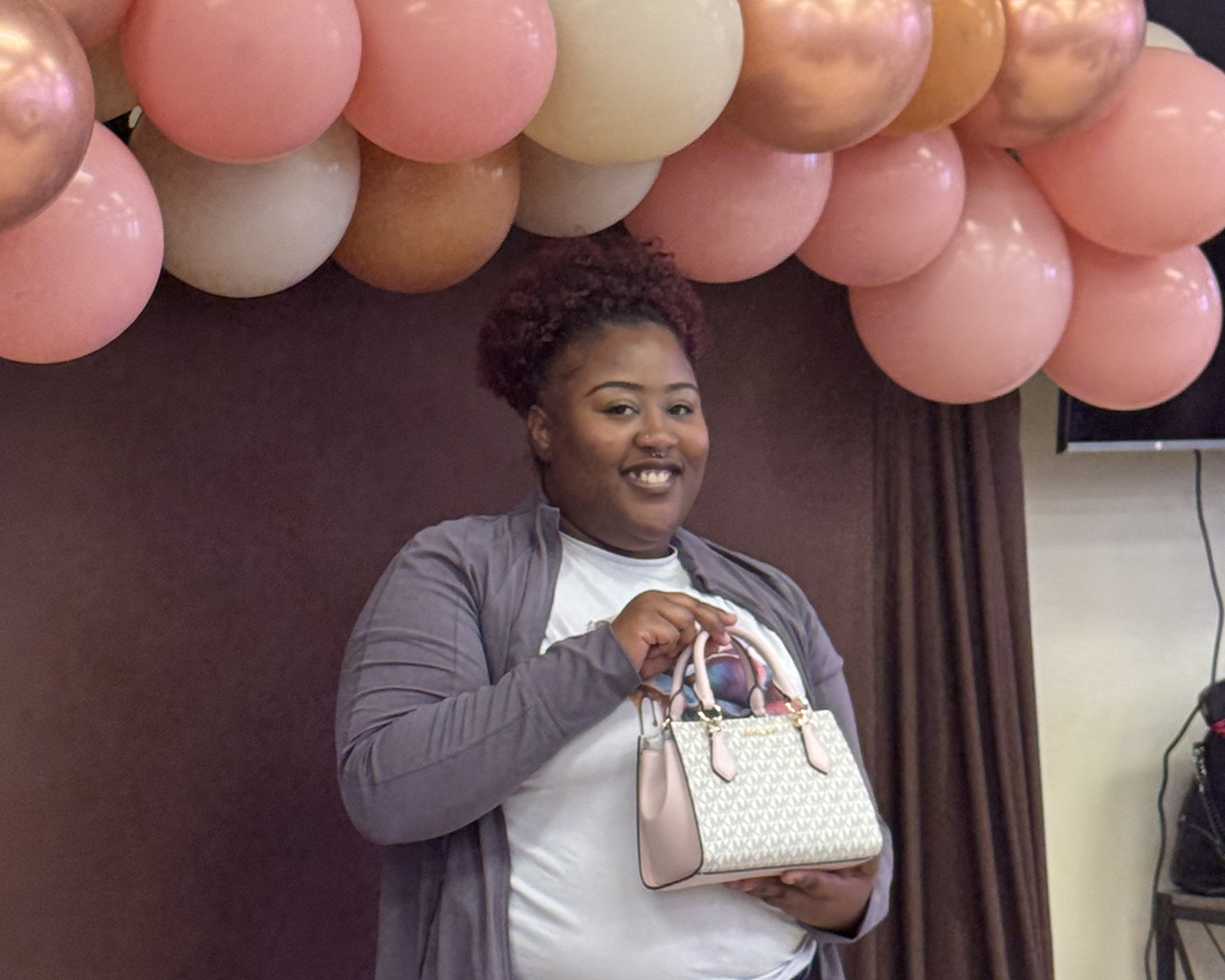 A smiling woman with curly hair holds a beige patterned purse in front of a backdrop of pink and beige balloons.