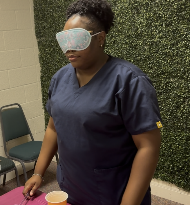 A woman wearing navy scrubs and a floral sleep mask over her eyes standing next to a pink table with a plastic cup, in a room with beige block walls and a green leafy decorated wall.