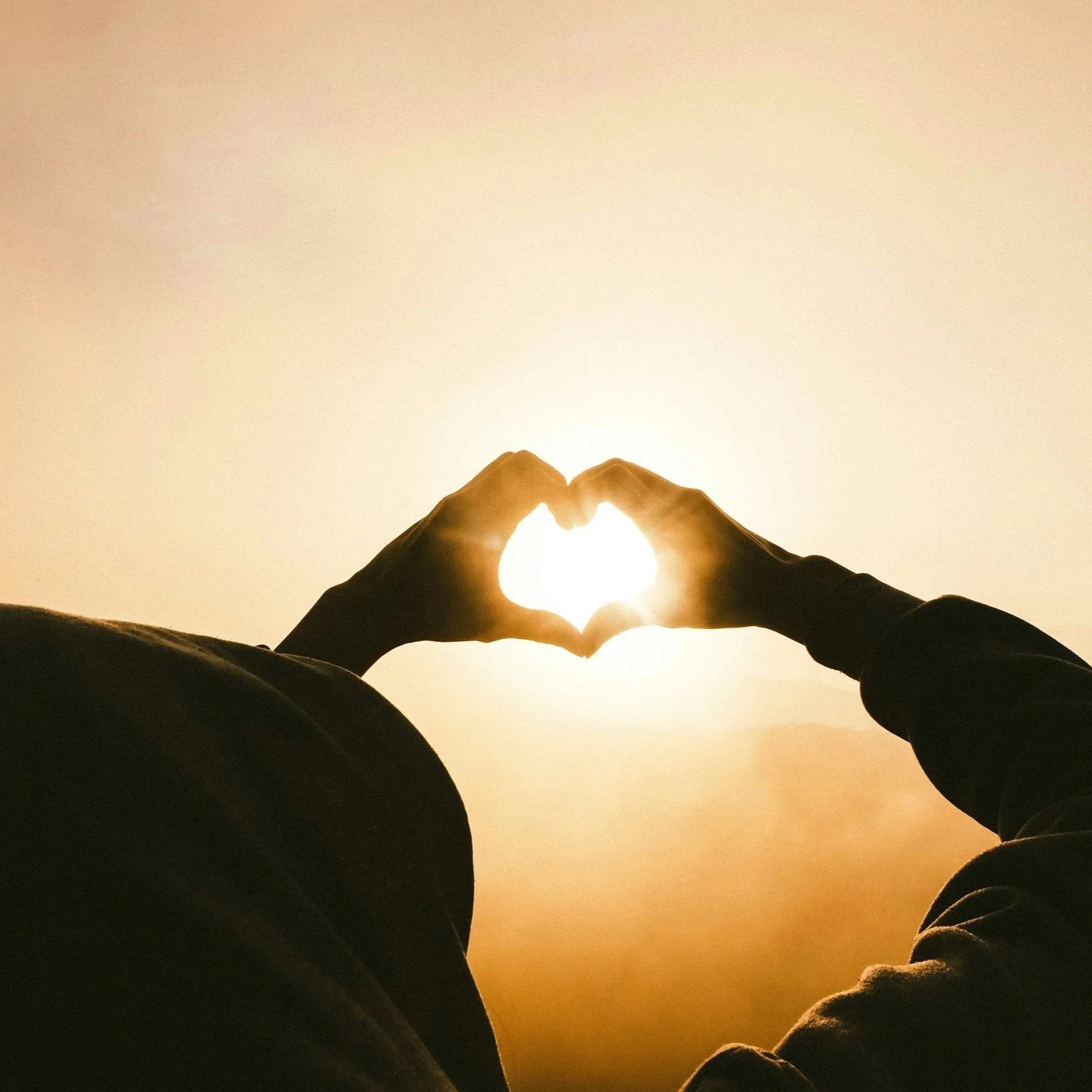 Person's silhouette forming a heart shape with hands around the sun during sunrise or sunset.