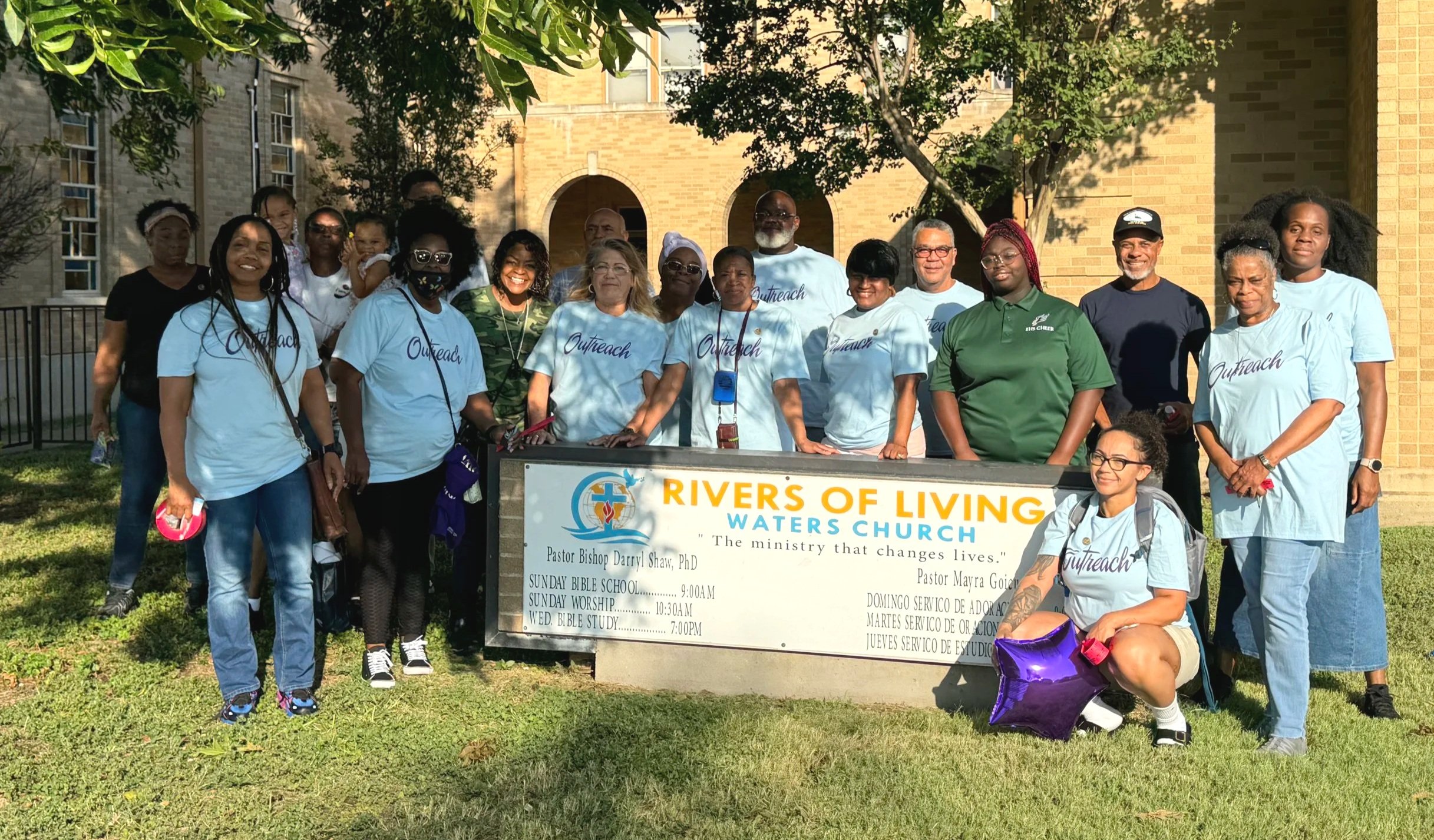 Group of diverse people standing outdoors around a sign for Rivers of Living Waters Church, with some wearing matching Outreach T-shirts, in front of a brick building and trees.