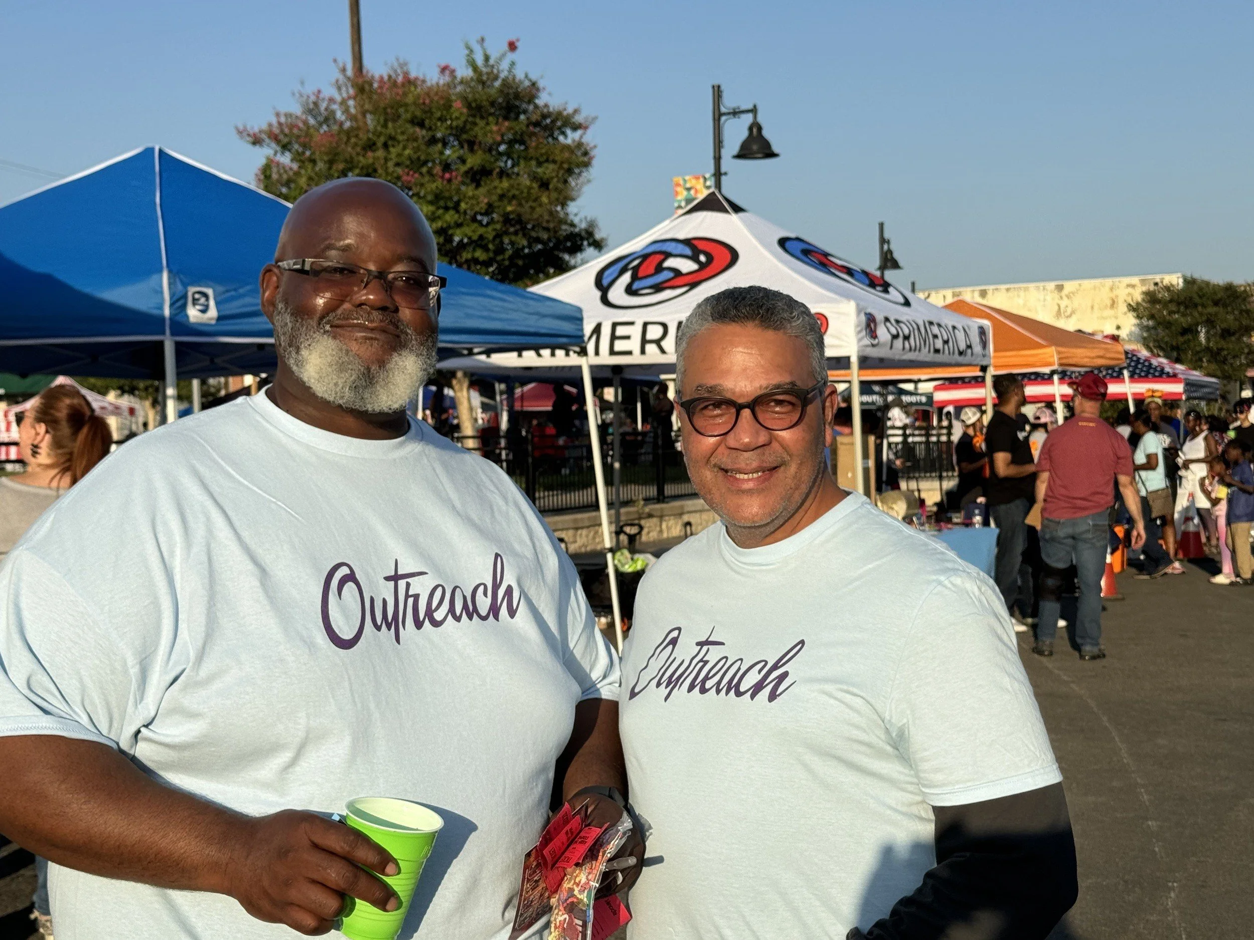 Two men wearing white 'Outreach' T-shirts standing outdoors at a festival, with tents and people in the background.