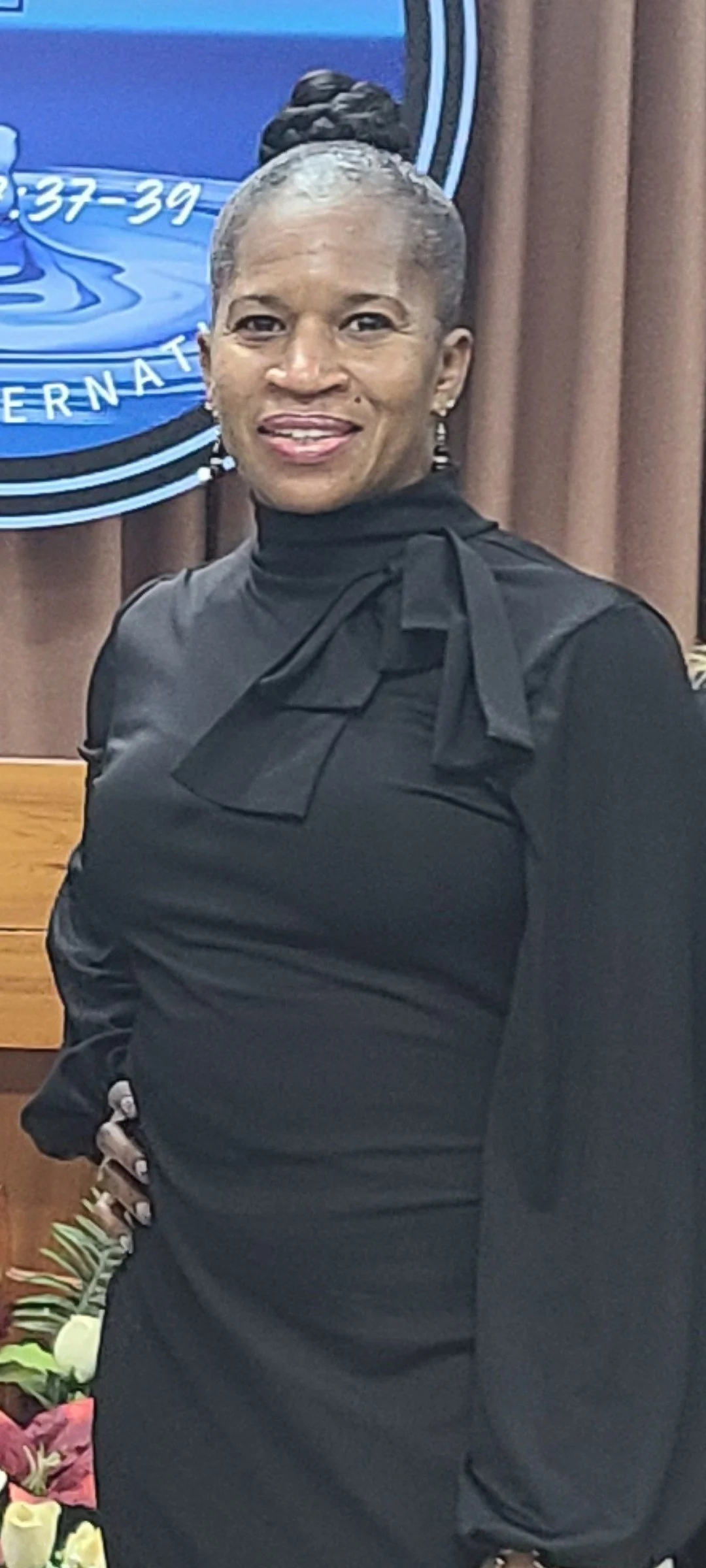 A woman with dark hair styled in an updo, wearing a black dress with a bow at the neck, standing indoors in front of a brown curtain and a blue sign that reads '37-39 INTERNATIONAL.'