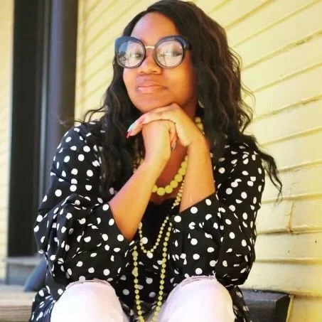 Woman with long wavy hair wearing black glasses, polka dot top, and yellow necklace, sitting outdoors with a yellow wooden wall in the background.
