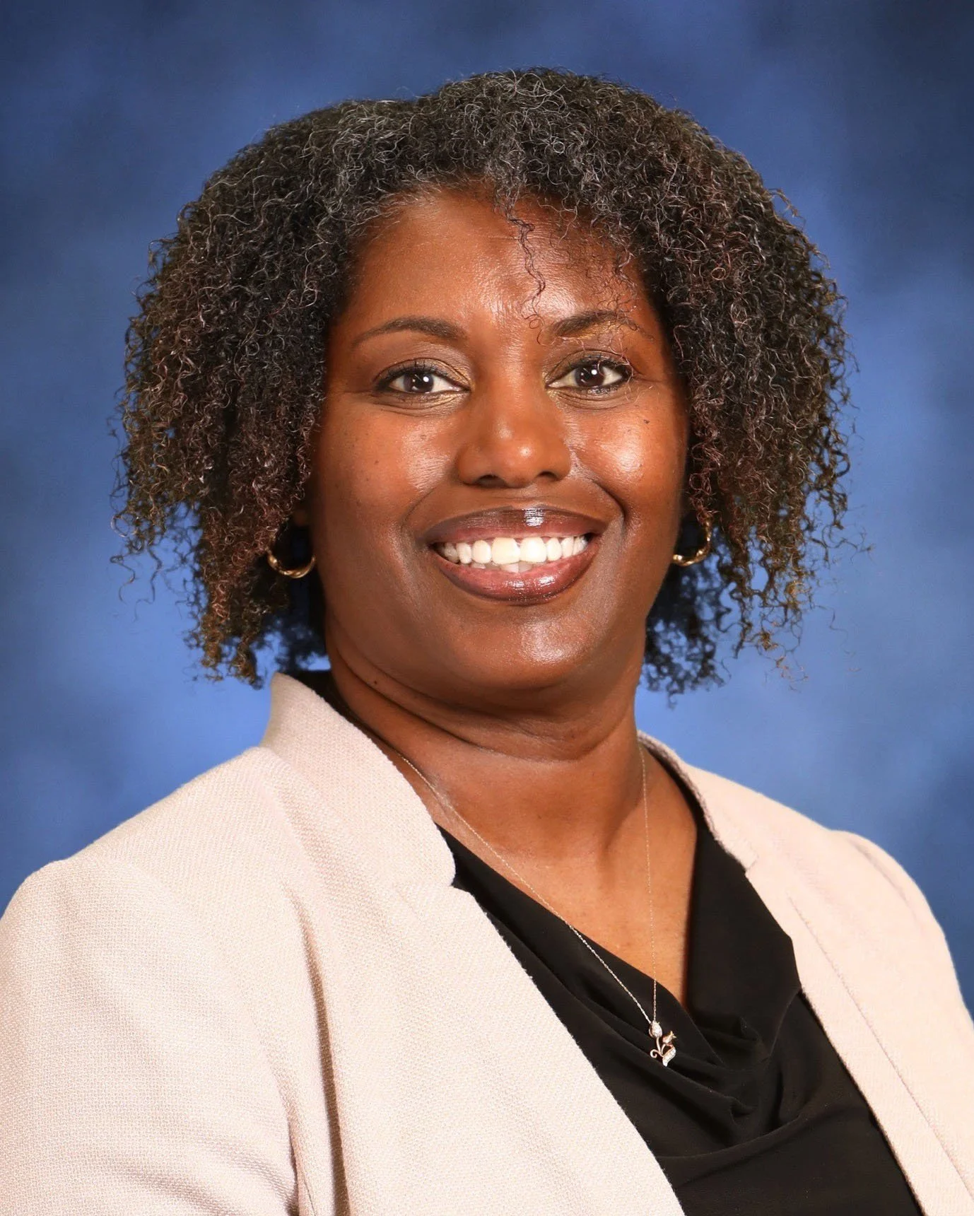 A professional headshot of a smiling woman with curly hair, wearing a cream blazer over a black top, against a blue background.