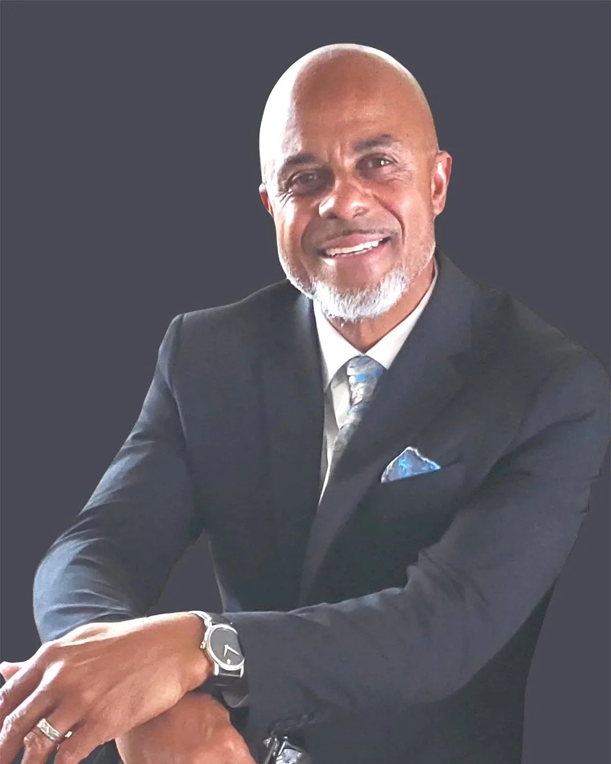 A professional man with a beard and glasses, wearing a dark suit, white shirt, patterned tie, and a wristwatch, seated against a plain dark background.