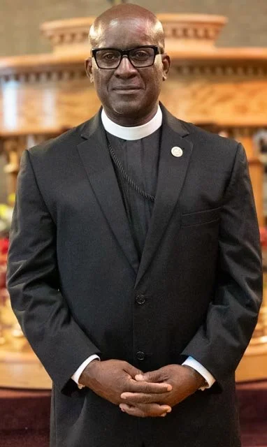 A man in a priest's black suit and clerical collar standing inside a church with wooden decor and a decorative arch in the background.