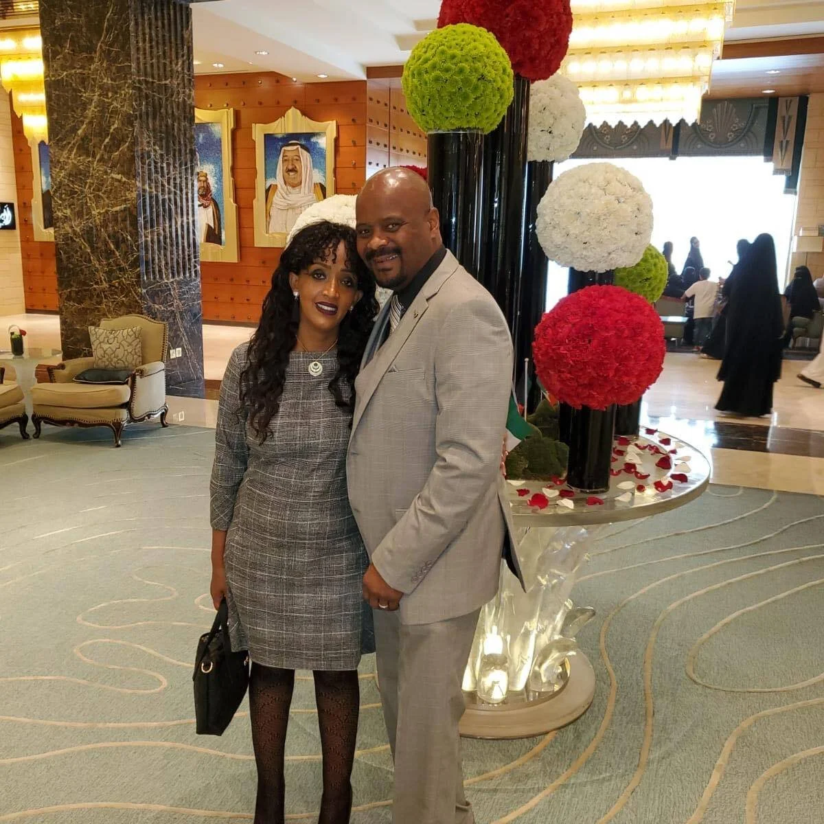 A couple dressed in formal attire standing in a hotel lobby with floral decorations and portraits on the wall.
