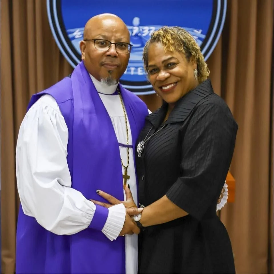 A priest and a woman are standing together indoors, smiling and holding hands. The priest is wearing a purple and white robe with a cross necklace, and behind them is a circular emblem or logo on the wall.