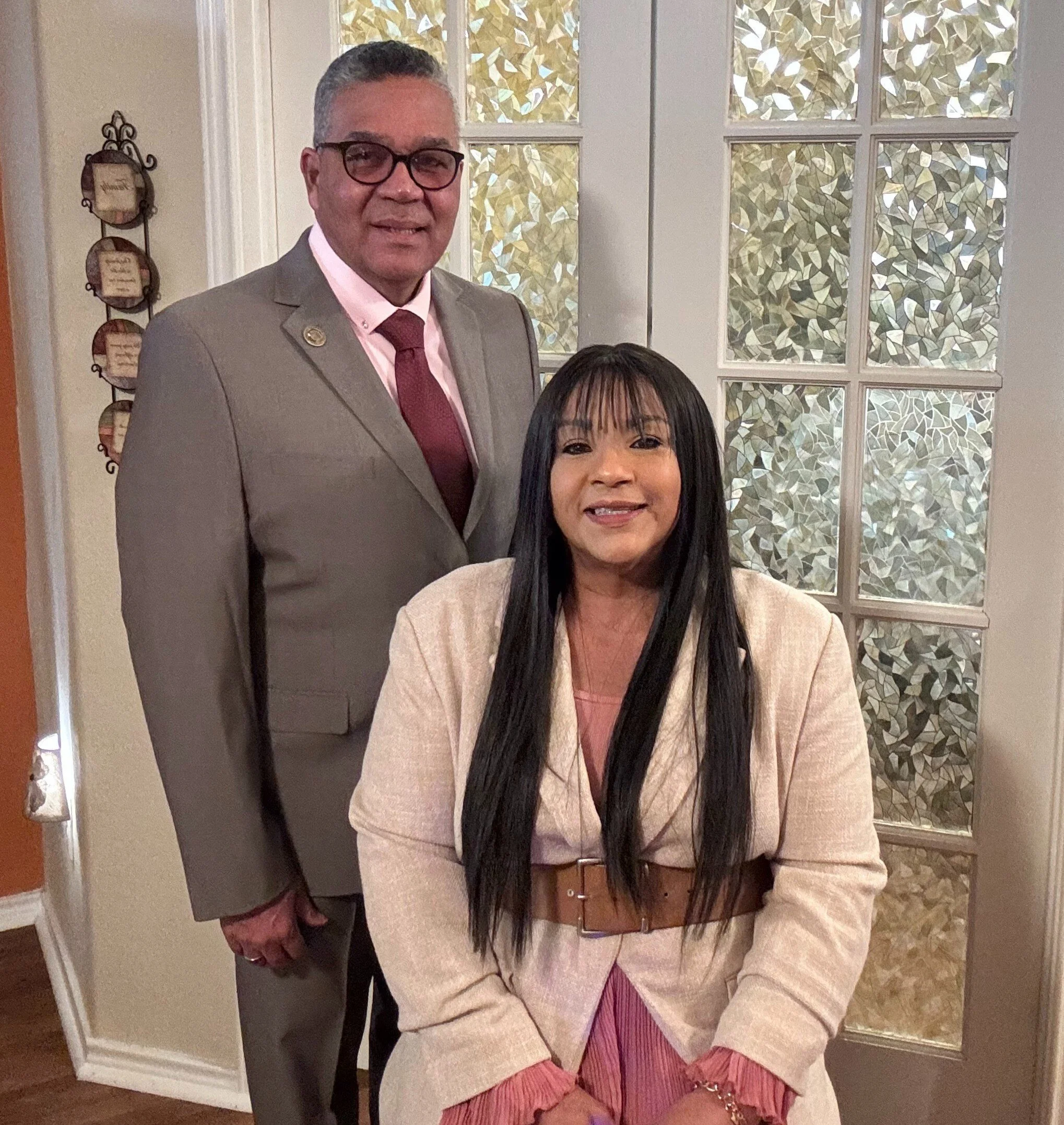A man in a gray suit and glasses standing behind a seated woman with long black hair, wearing a beige blazer and pink dress inside a room with decorative stained glass windows and wall decorations.