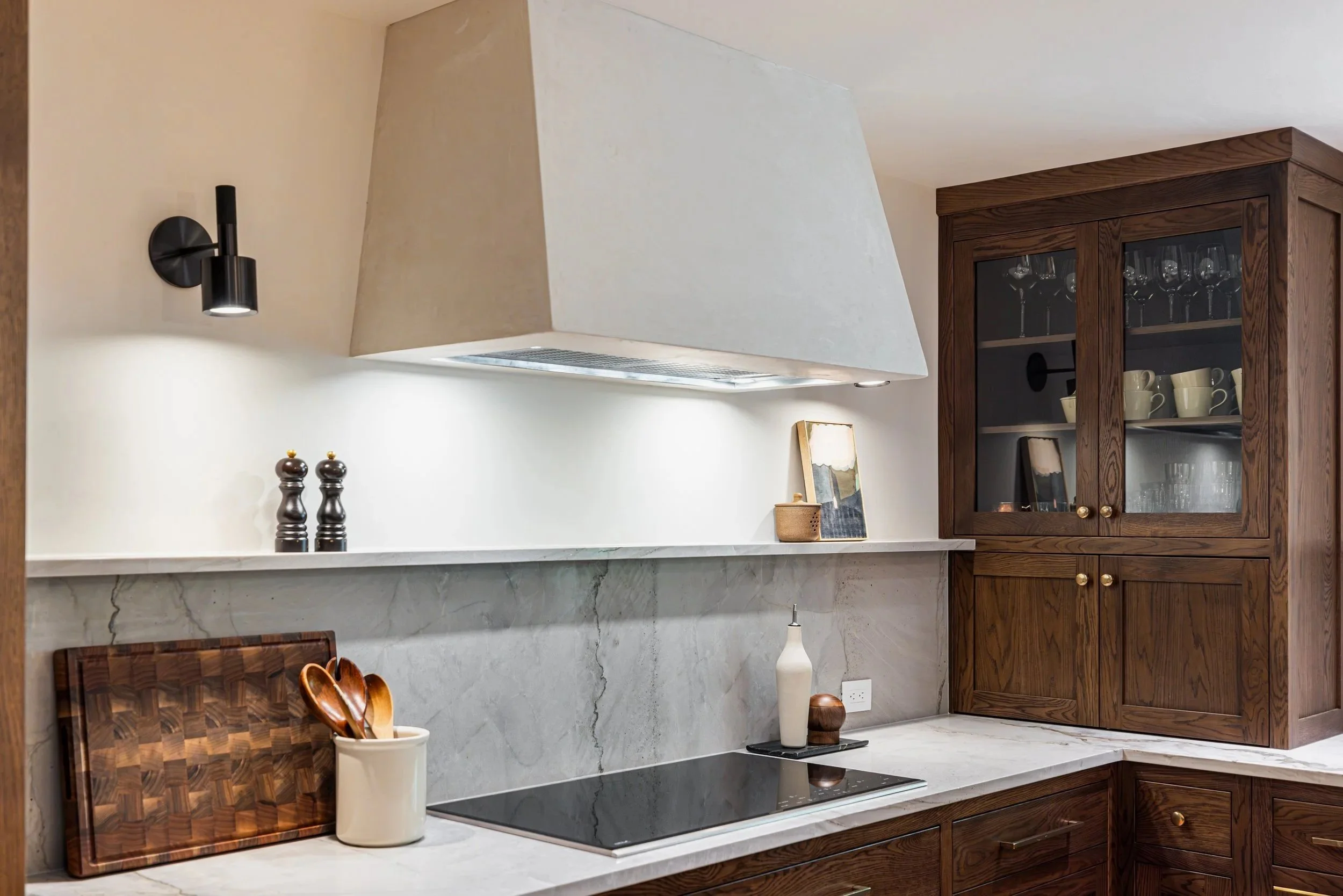 Kitchen with wooden cabinets, marble countertop, black stove, and a white vent hood. Decorative items include two black pepper mills, a framed artwork, a small basket, and a ceramic container holding wooden spoons. There's also a black wall-mounted light fixture.