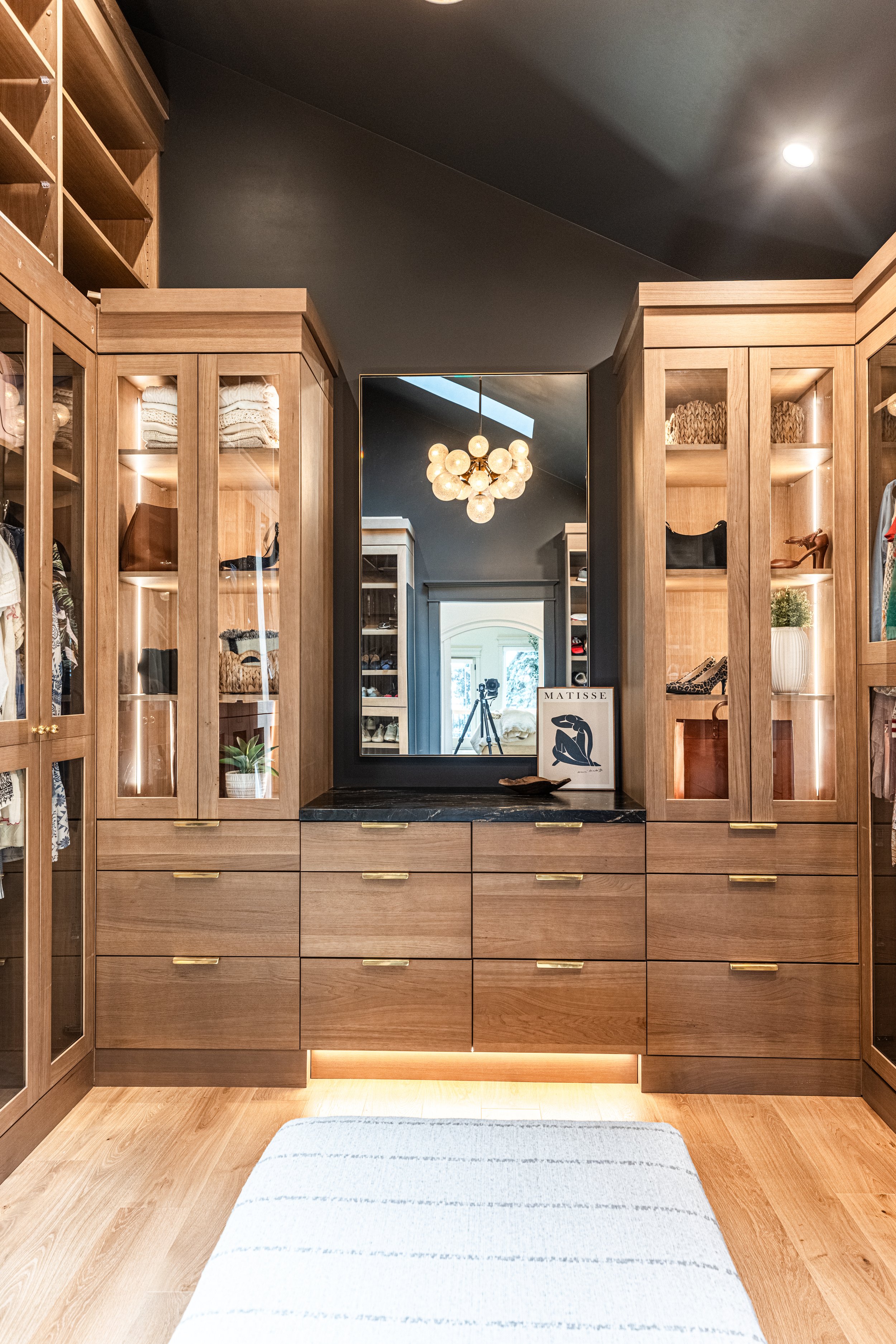 View of a well-organized walk-in closet with wooden cabinets and drawers, a large mirror with a black frame, a modern chandelier hanging from a dark ceiling, and a light-colored rug on a wooden floor.
