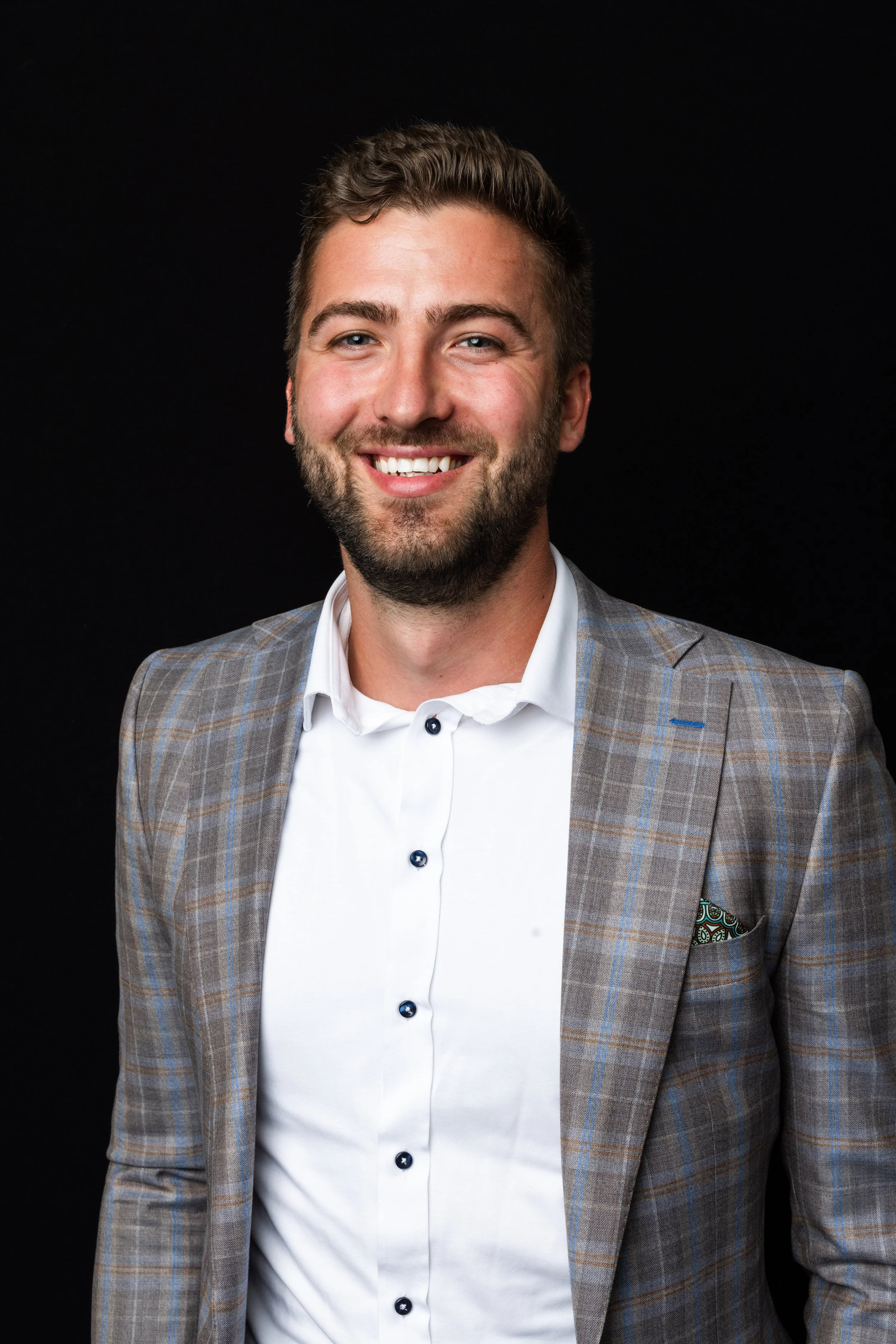 A smiling man with a beard and short brown hair wearing a light gray plaid blazer over a white shirt against a black background.