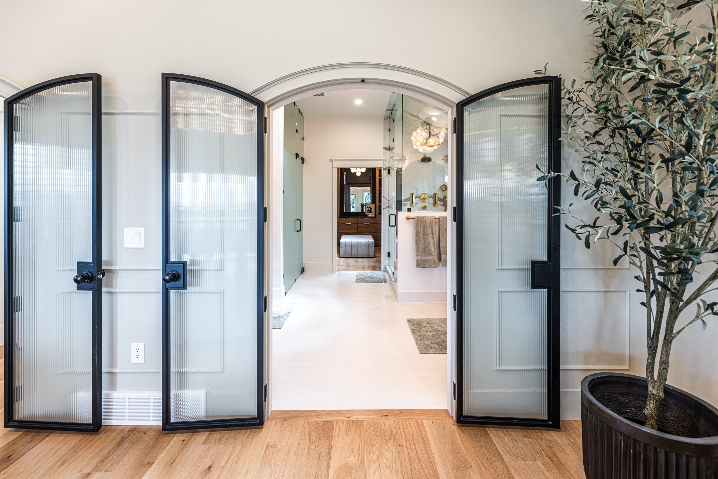 Interior view through glass doors revealing a hallway with modern lighting, a vanity area, and a potted tree on the right.
