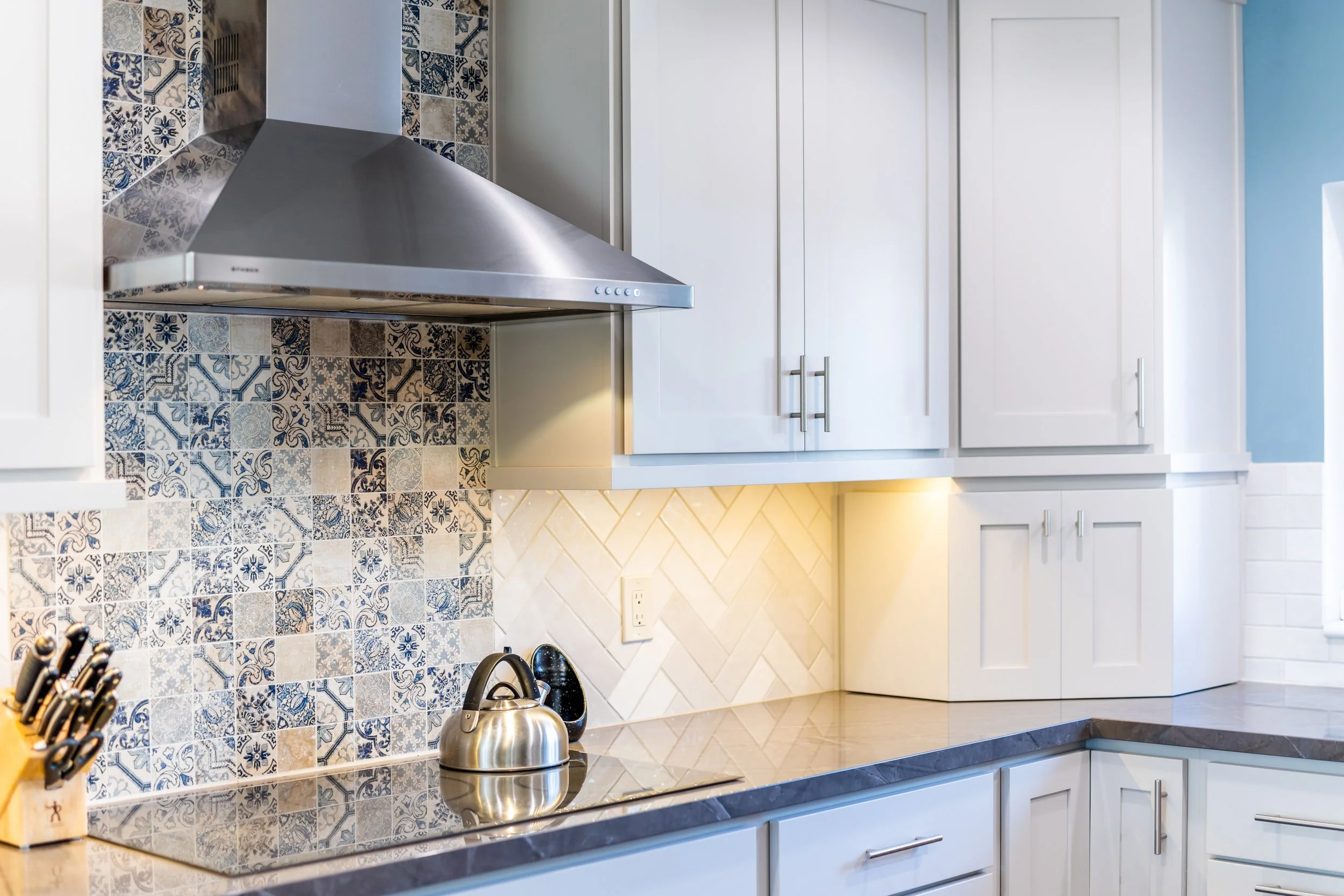 Modern kitchen with white cabinets, patterned tile backsplash, stainless steel range hood, and a stainless steel kettle on a dark granite countertop.