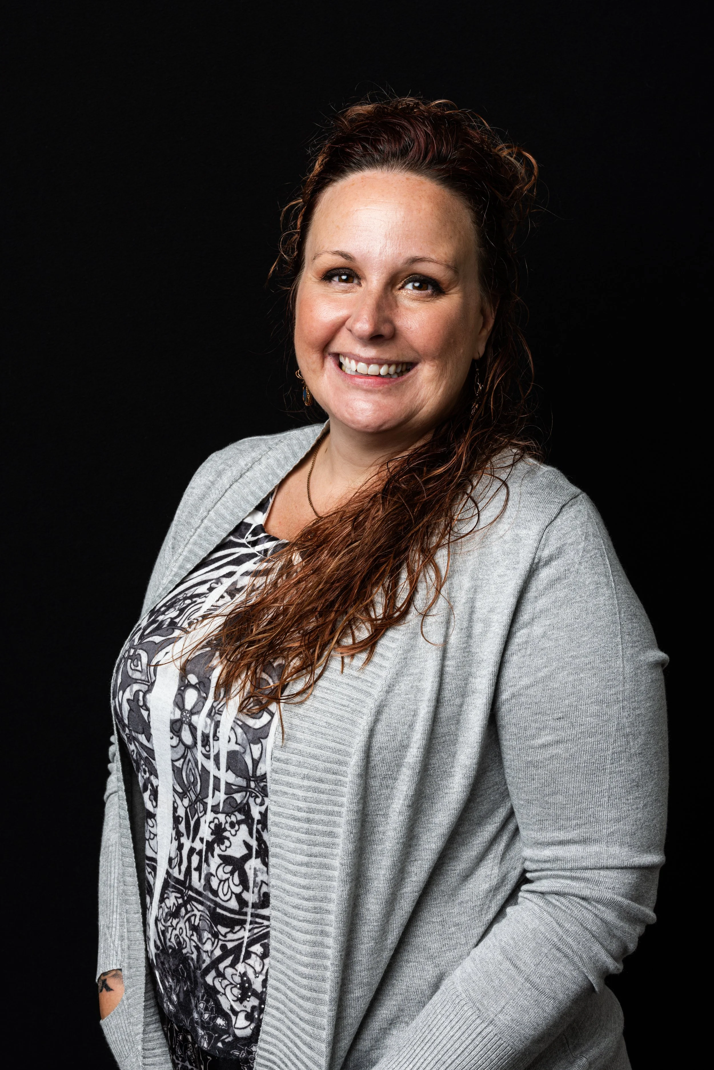 A woman with long, wavy auburn hair, smiling, wearing a gray cardigan over a black and white patterned blouse, standing against a black background.