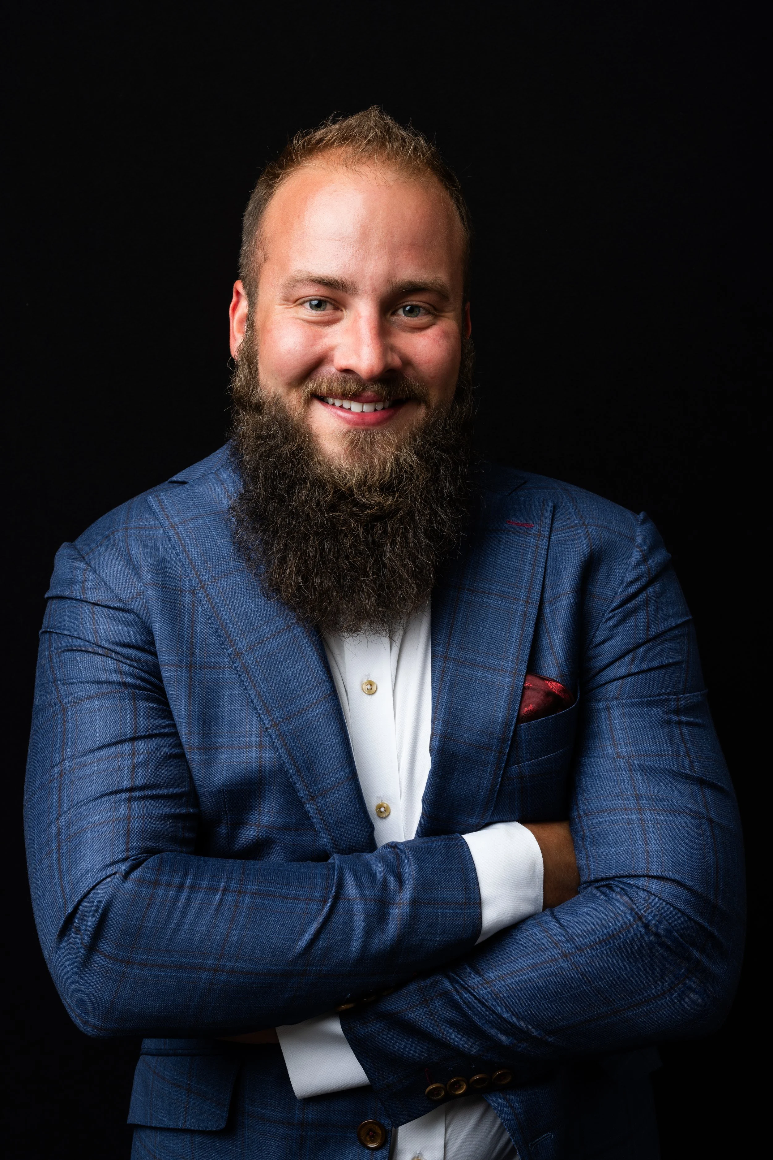 Portrait of a smiling man with a beard, wearing a blue checkered suit jacket, white shirt, and red pocket square, with arms crossed against a black background.