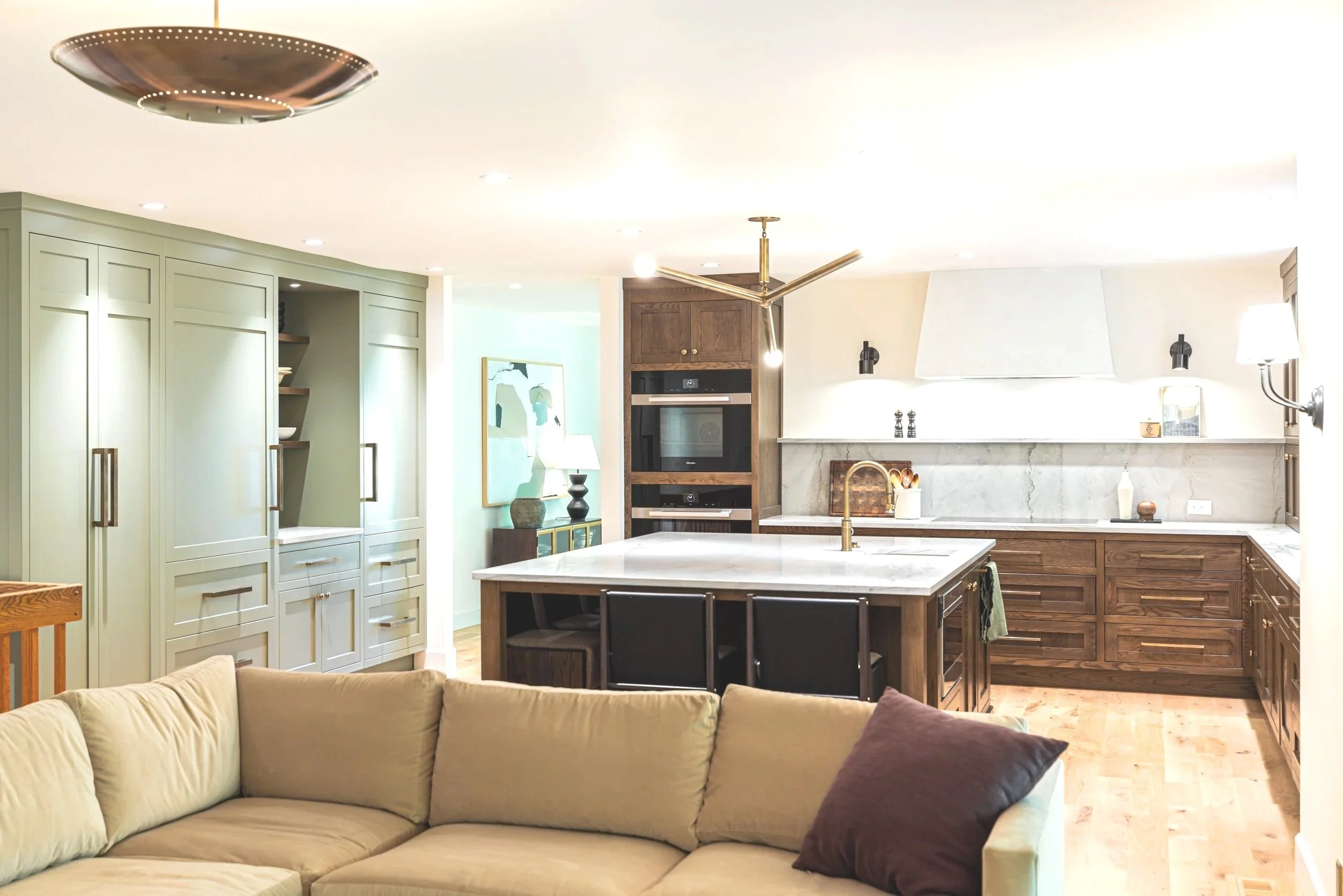 Modern kitchen with beige and brown cabinets, a white marble island, and a stove with a white range hood. There is a beige curved sofa in the foreground.