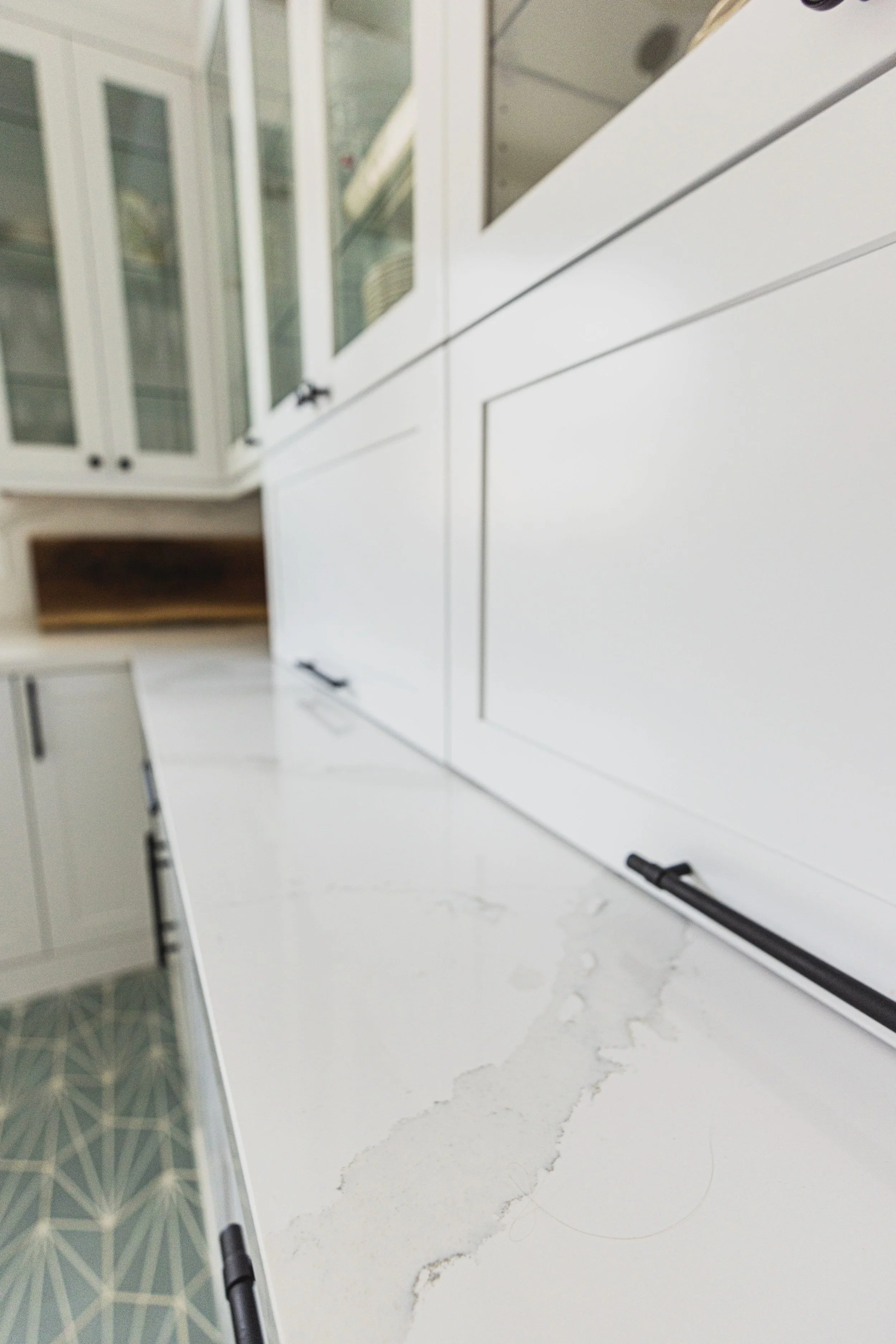 White kitchen cabinets with glass doors and black handles, white marble countertop with grey veining, and a section of a tiled floor with a geometric pattern, possibly near a kitchen or pantry area.