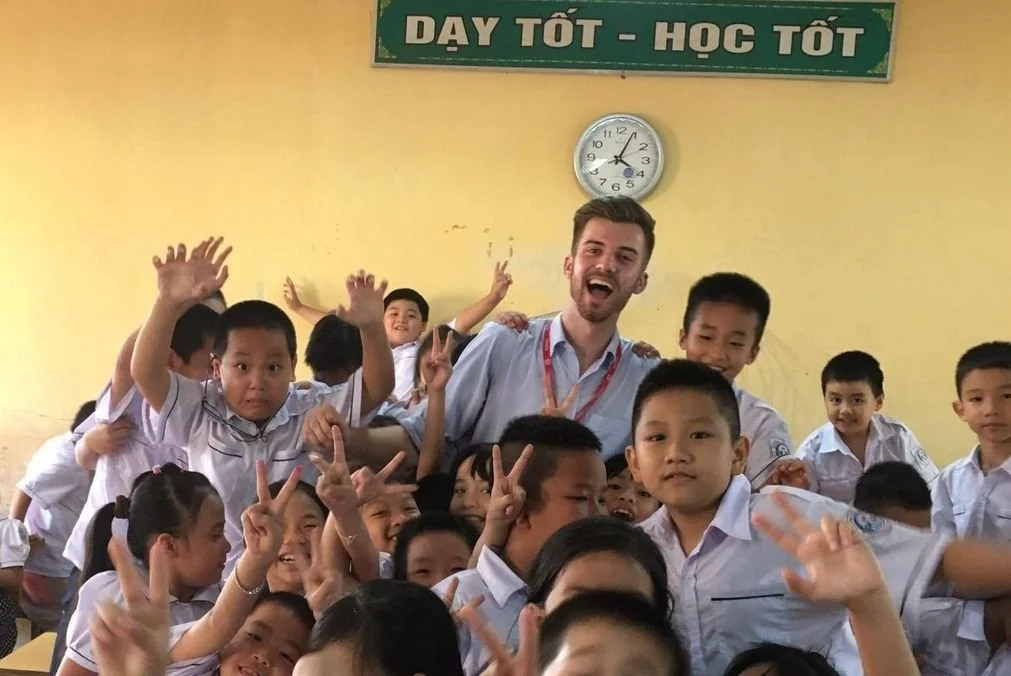 A group of elementary school students in white uniforms posing with a smiling teacher or adult in a classroom. The students are making peace signs and waving. There is a green sign with Vietnamese text and a clock on the yellow wall behind them.