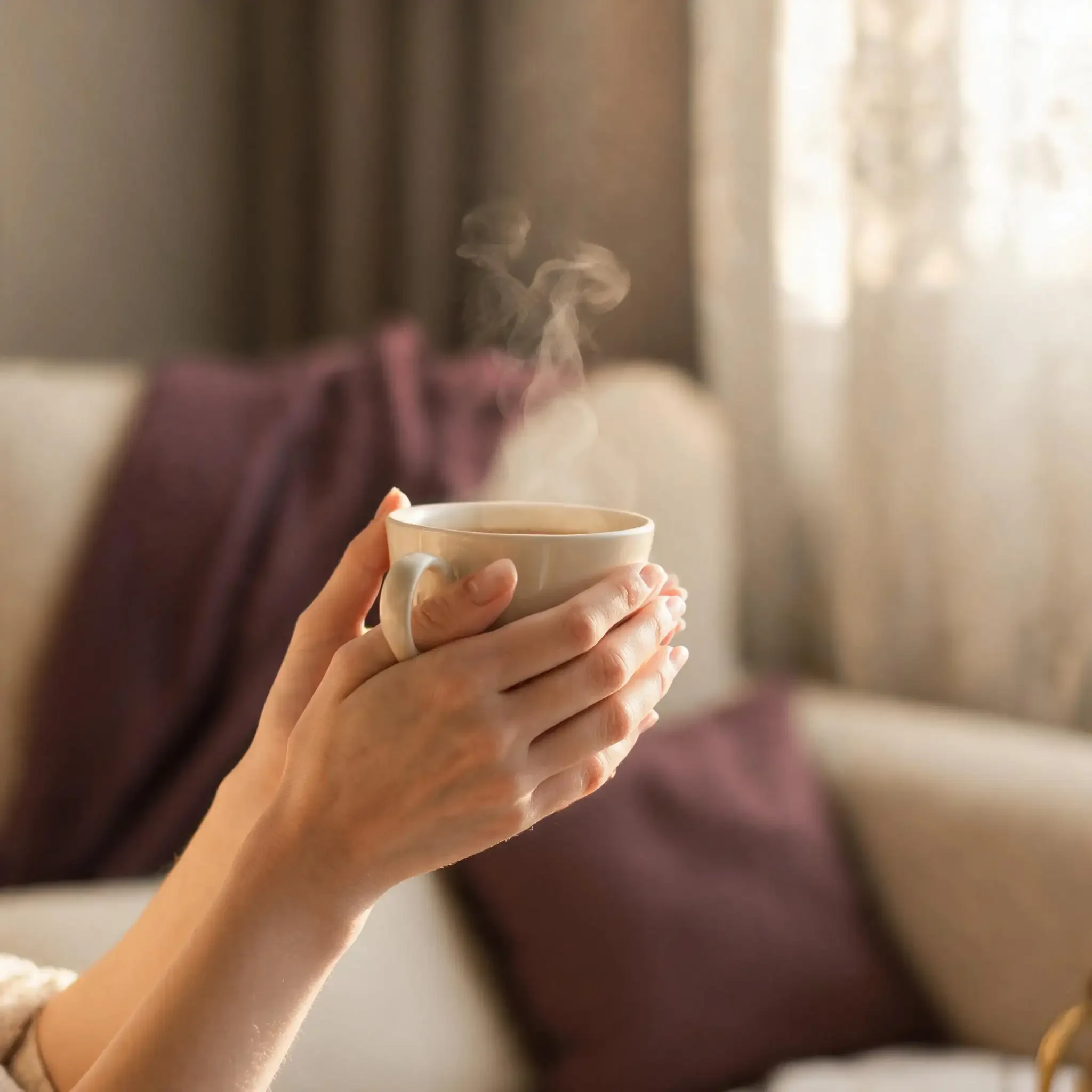 Close-up of someone holding a steaming cup of coffee in front of a blurred background with sunlight streaming in.